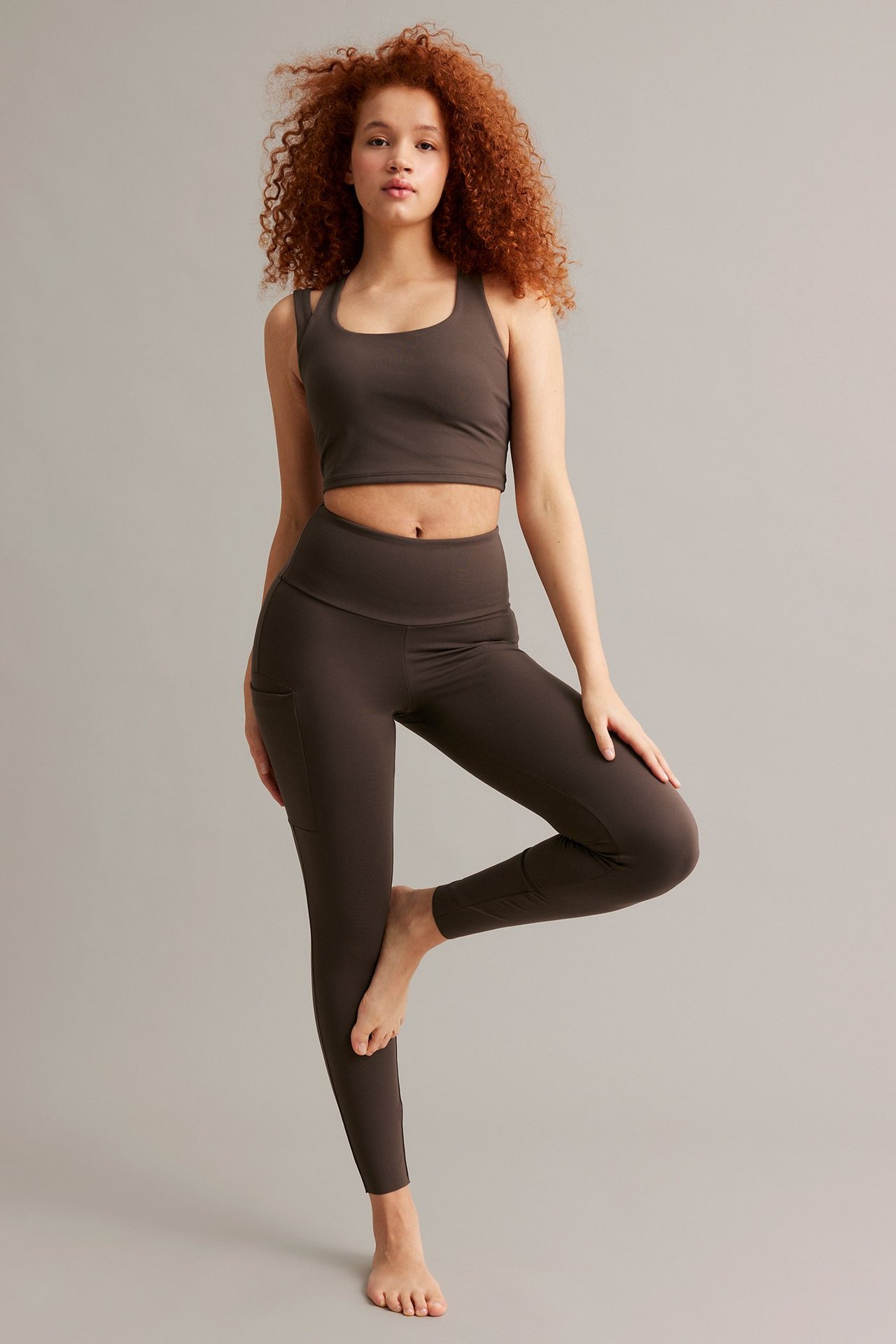 Forfra, kvinne i en tettsittende brun sportstopp og matchende brune leggings med sidelomme.