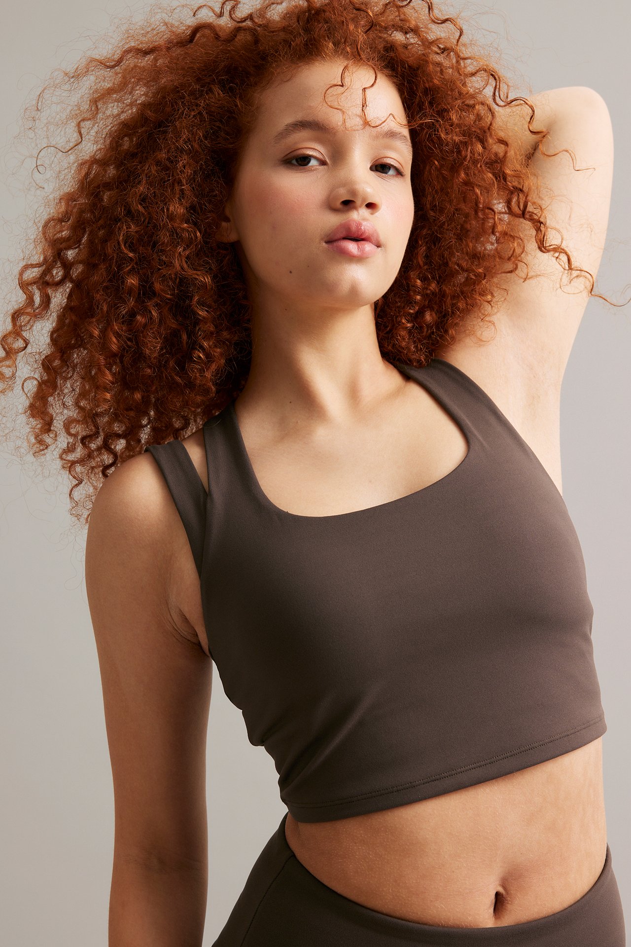 Nainen edestäpäin tummanruskeassa, pyöreäkauluksisessa sportti-croptopissa, leveillä olkaimilla ja yhteensopivilla alaosilla.
