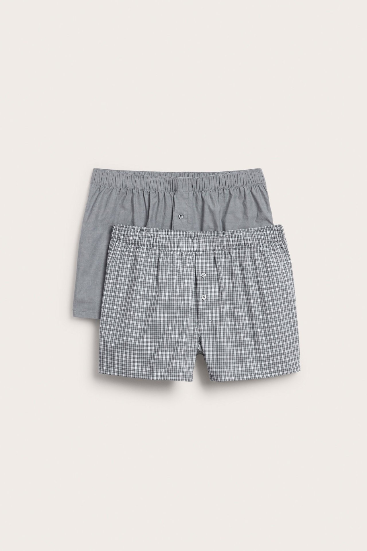 Frontvy av två par vävda boxerkalsonger i bomull: ett grårutigt, ett enfärgat grått, båda med knappgylf.