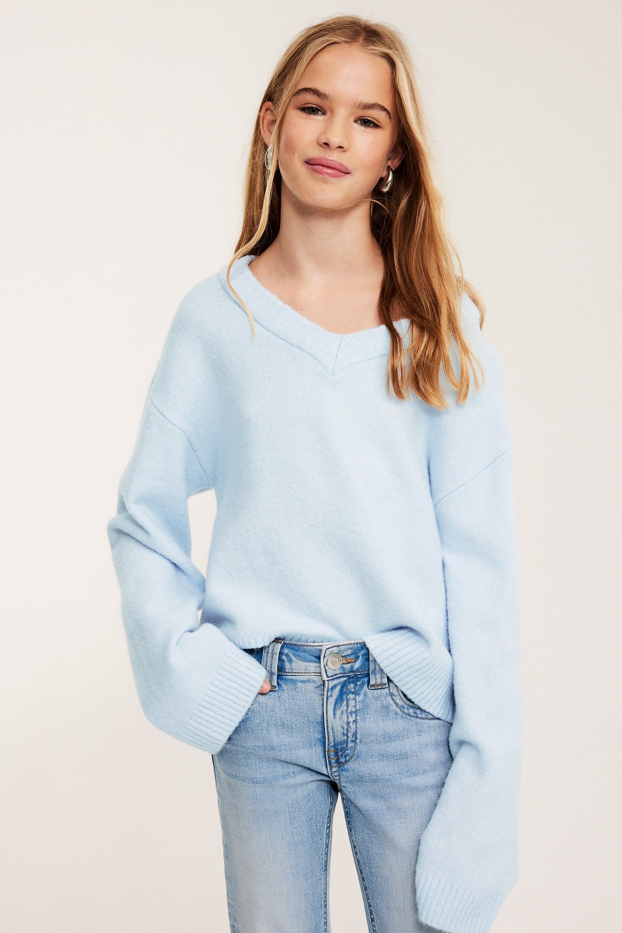 Frontbilde av et barn i lyseblå v-halset strikkegenser, lyse jeans og ringøreringer.