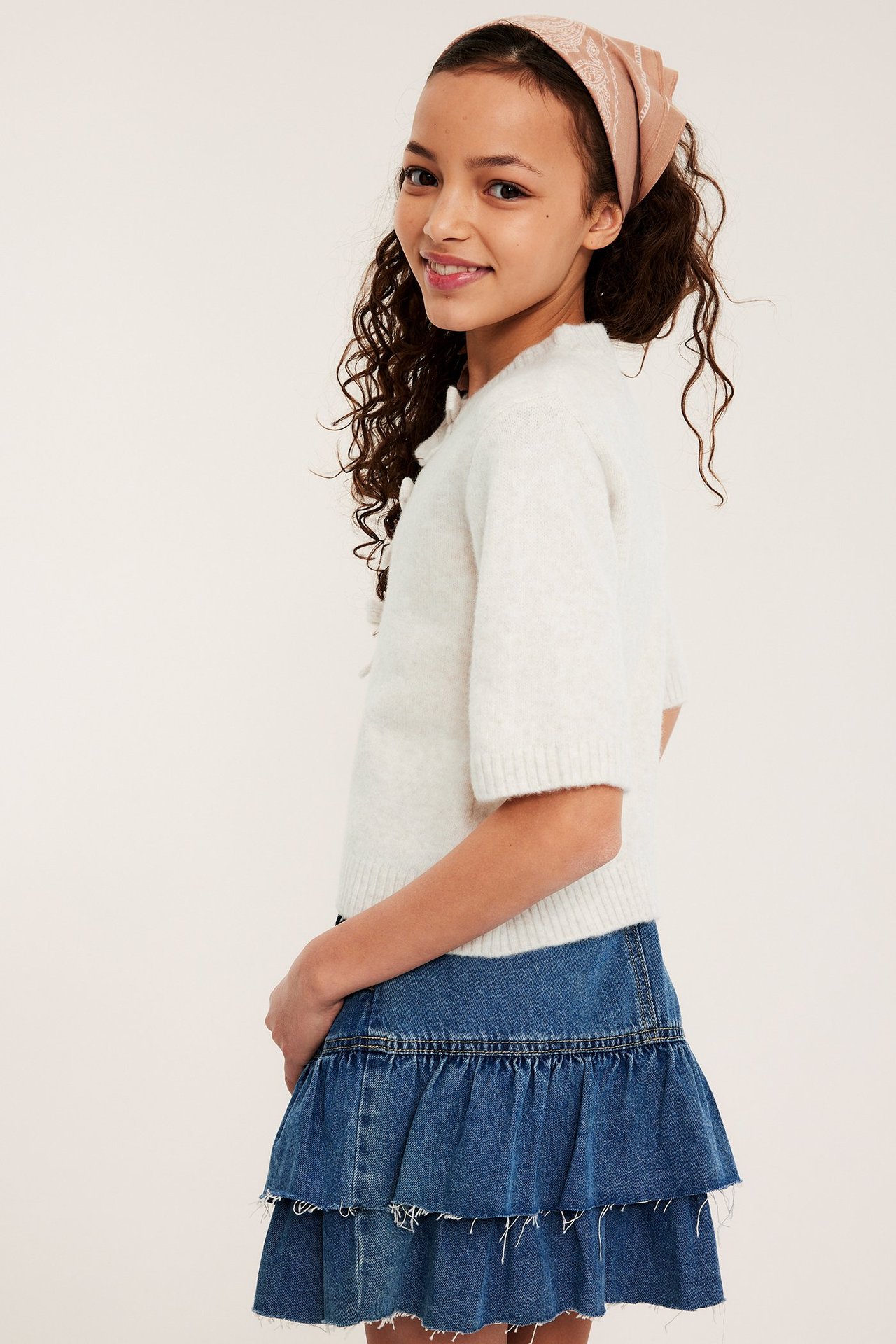 Widok z boku: Dziecko w białym melanżowym sweterku z kokardkami, niebieskiej jeansowej spódniczce i różowej wzorzystej bandanie.