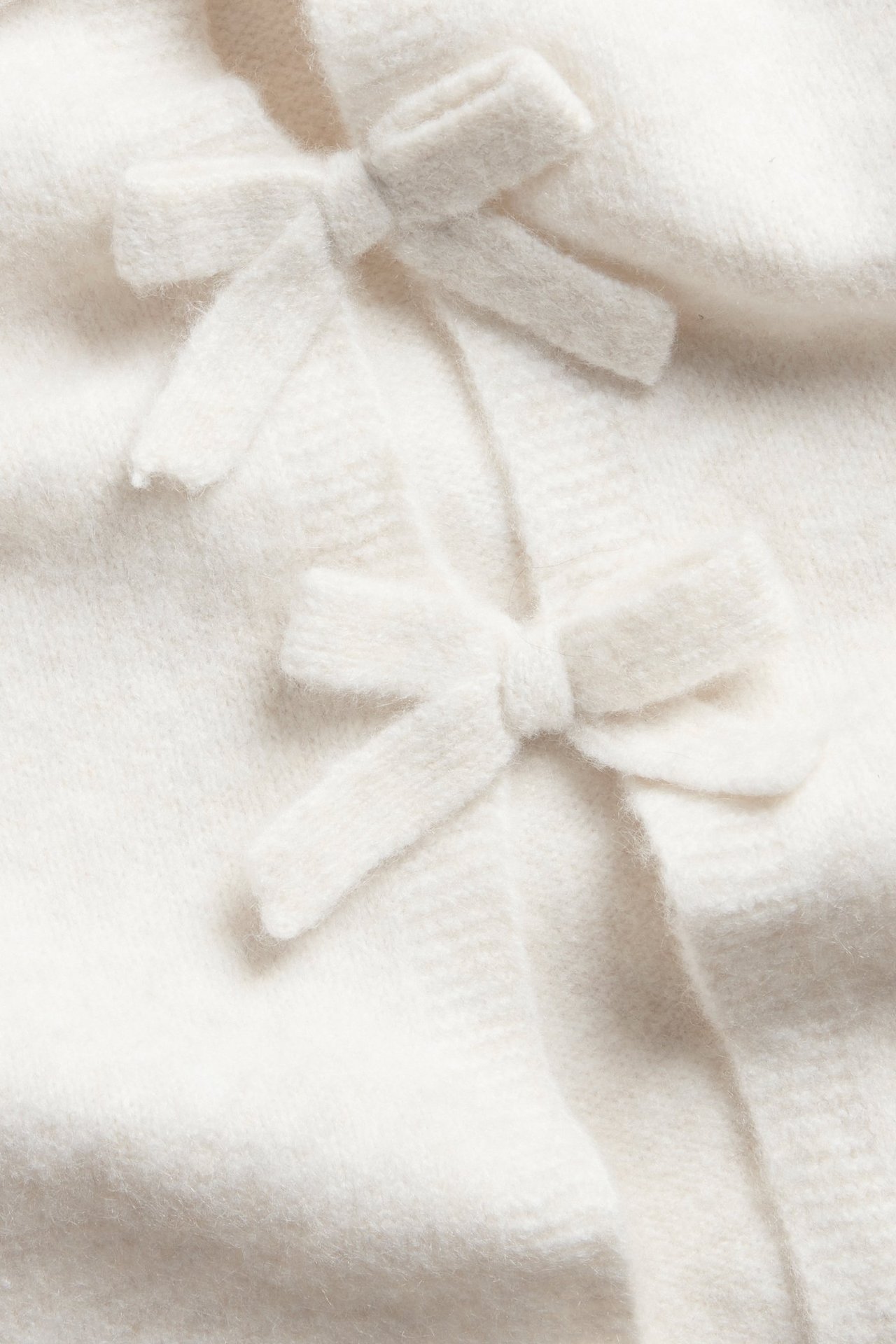 Zbliżenie na biały, drobno tkany, krótki kardigan z dwiema kokardkami.