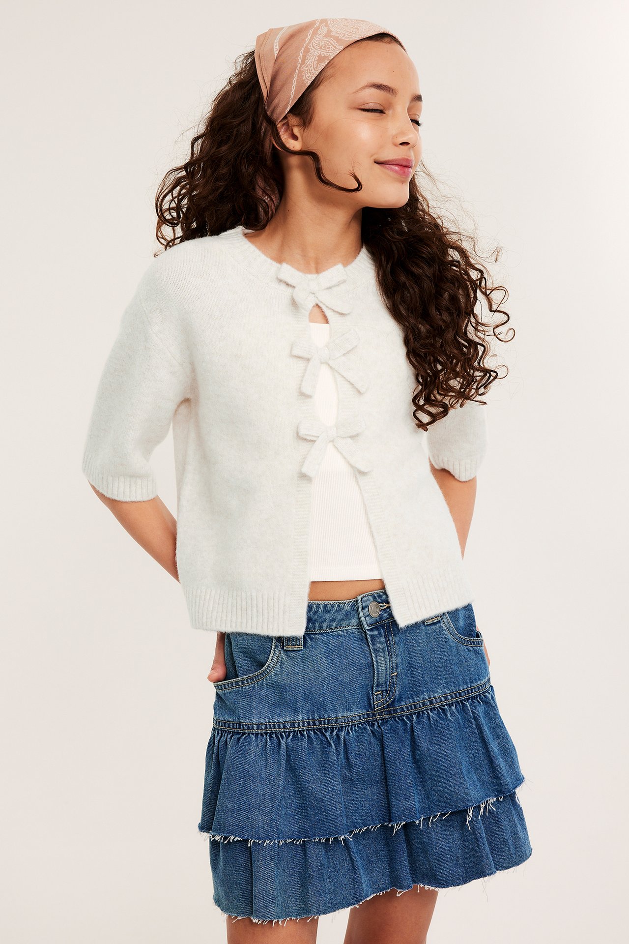 Widok z przodu: Dziecko w białym krótkim kardiganie z kokardami, topie, niebieskiej jeansowej spódniczce, beżowej bandanie.