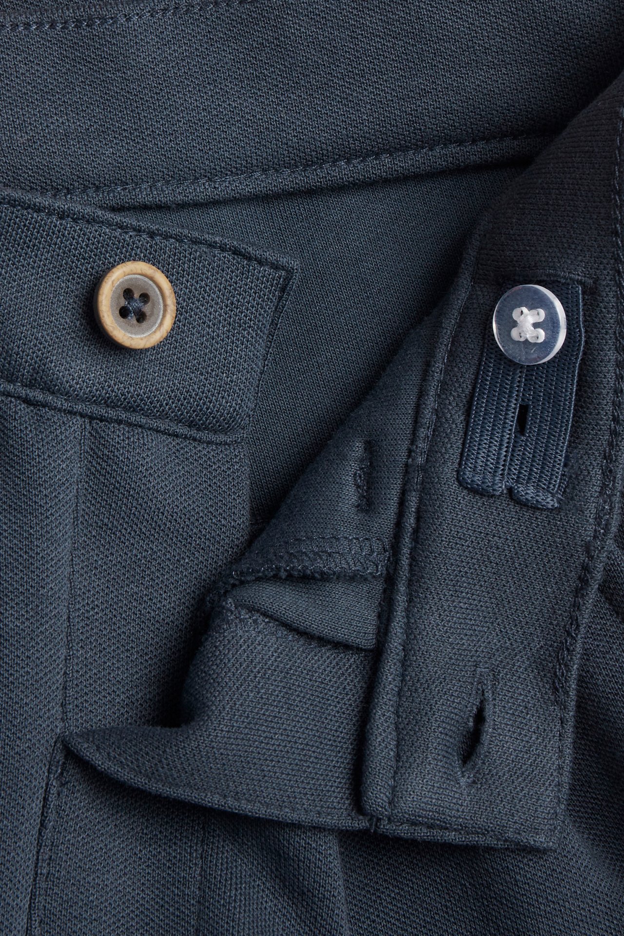 Lähikuva tummansinisistä lasten housuista, joissa edessä nappi ja säädettävä kuminauhavälikappale.