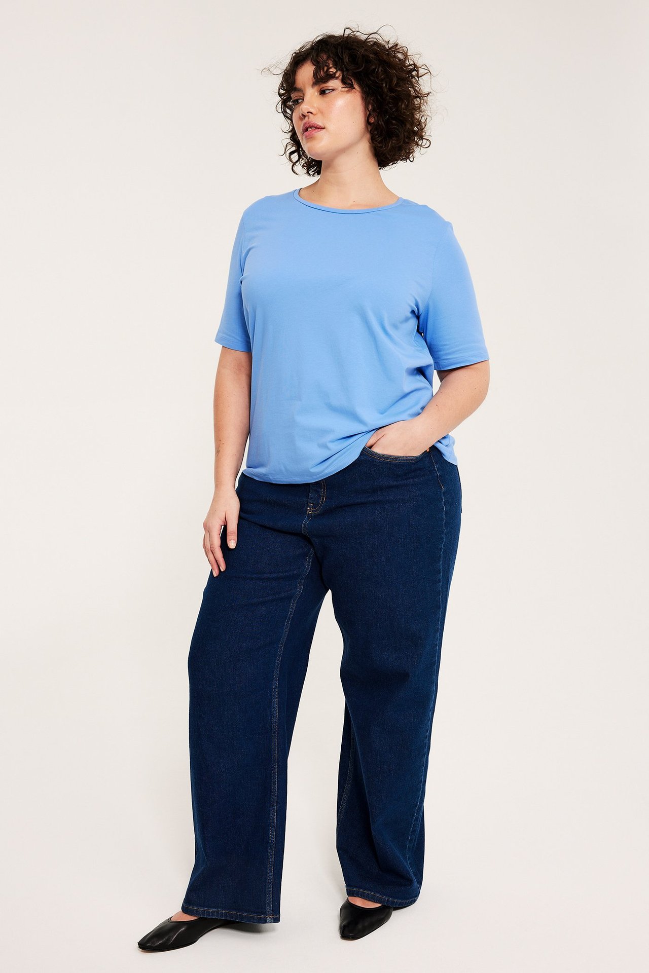 Frontbilde av kvinne i ensfarget lys blå bomulls-T-skjorte, mørkeblå jeans og svarte flate sko.
