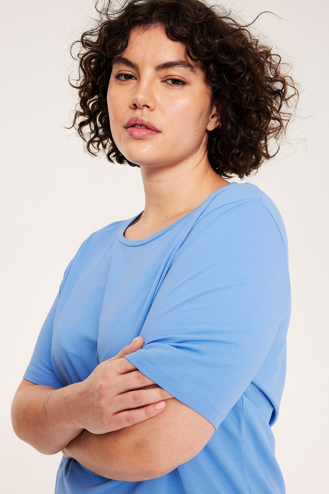 Nainen edestä ja sivulta, yllään vaaleansininen, yksivärinen puuvillainen t-paita, kädet ristissä.