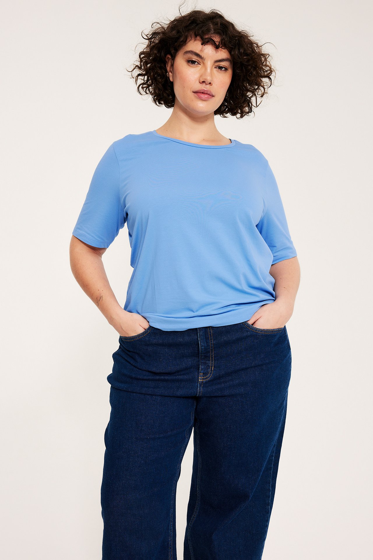 Frontalvisning: Kvinne i lys blå kortermet bomulls-t-skjorte og mørkeblå jeans.