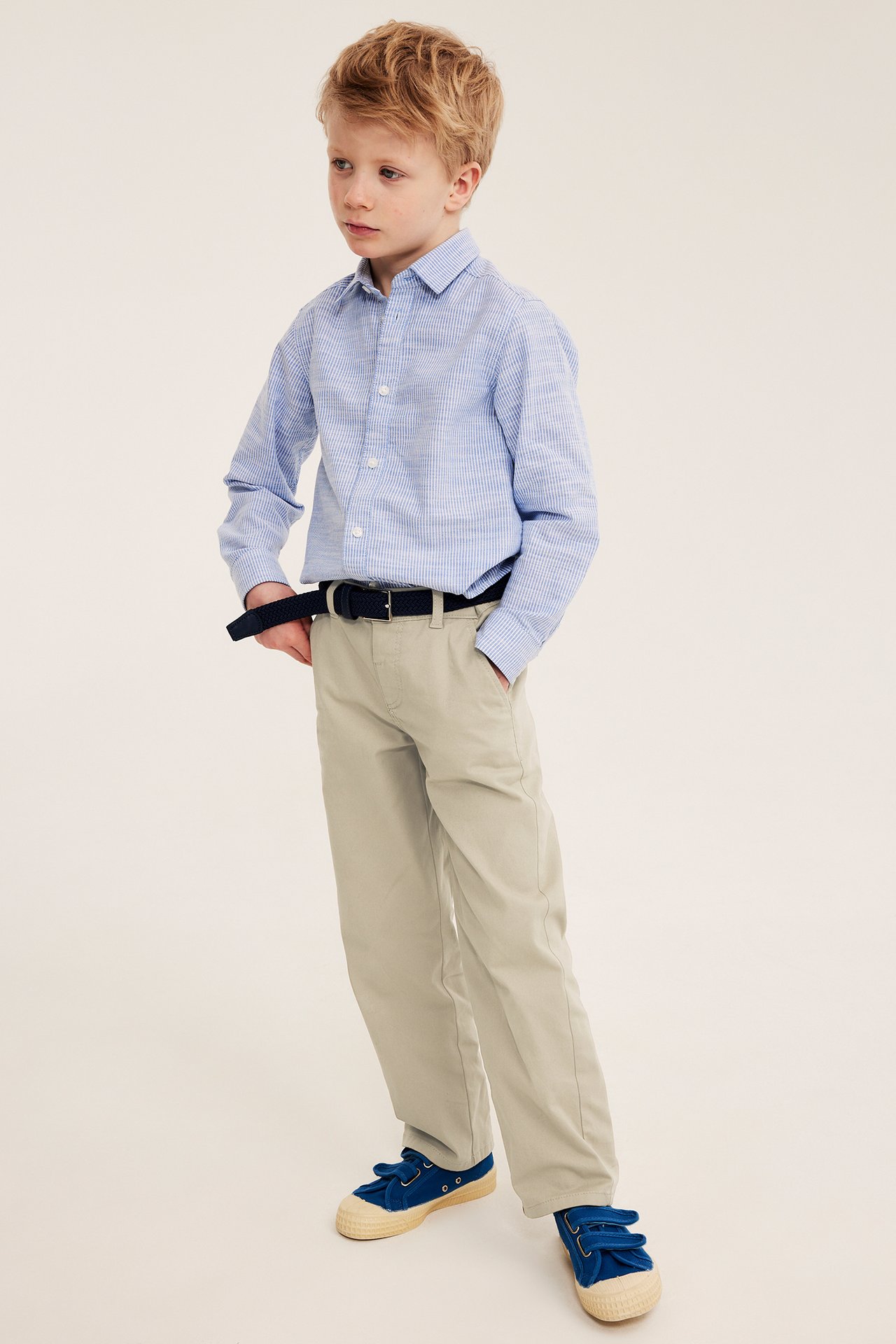 Framifrån: Ett barn i beige chinos, en blårutig skjorta, ett mörkblått bälte och blå sneakers.