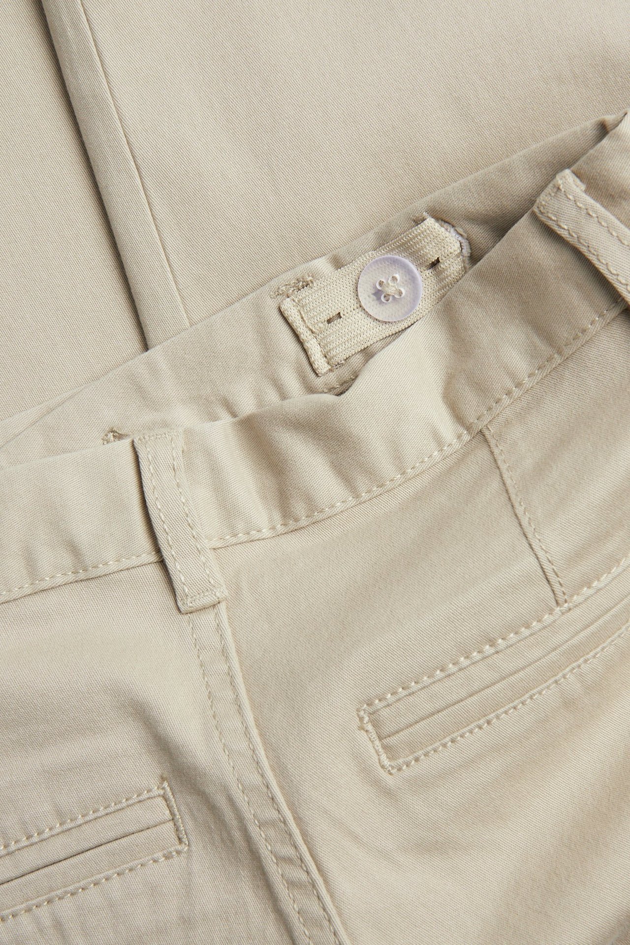Zbliżenie beżowych dziecięcych chinosów z bawełnianego twillu, pokazuje regulowany wewnętrzny pas z guzikiem i detale tylnych kieszeni.