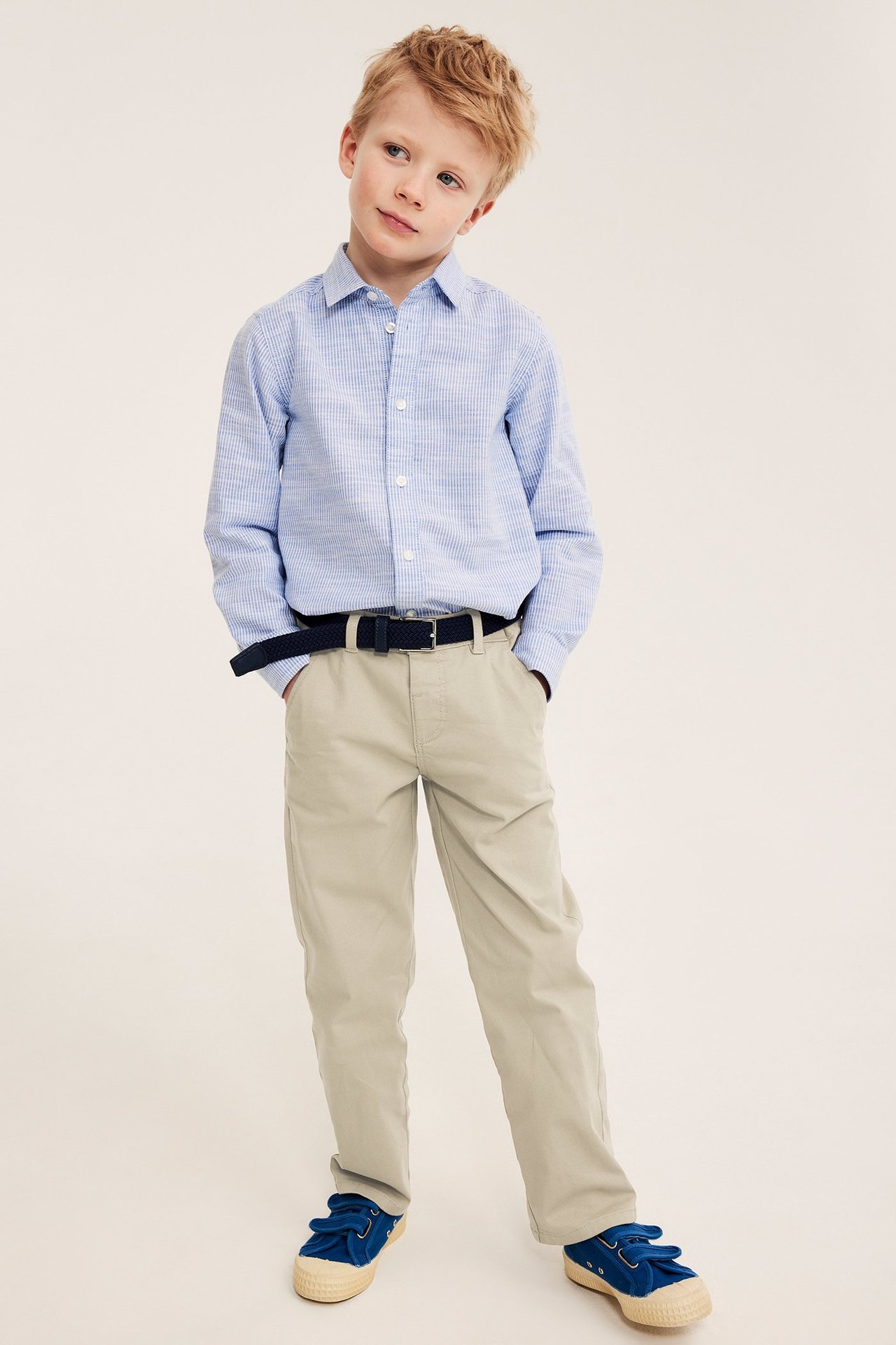 Z przodu, dziecko w beżowych chinosach, granatowym pasku, jasnoniebieskiej koszuli w kratę i niebieskich butach.