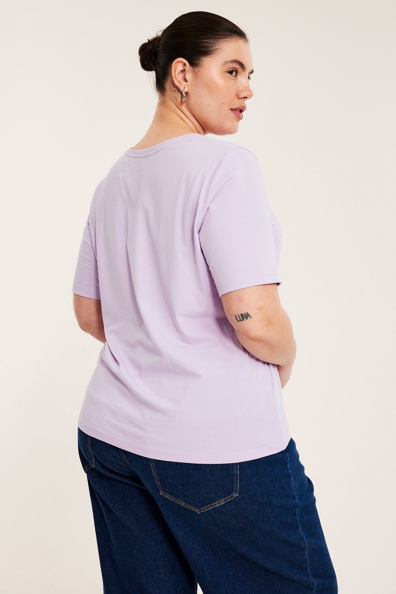 Widok z tyłu kobiety w jednolitej, jasnofioletowej koszulce i ciemnoniebieskich dżinsach.