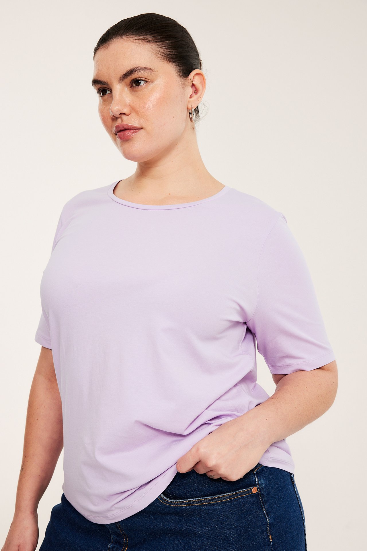 Widok z przodu: kobieta w jasnofioletowym t-shircie z okrągłym dekoltem, granatowych dżinsach i srebrnych kolczykach.