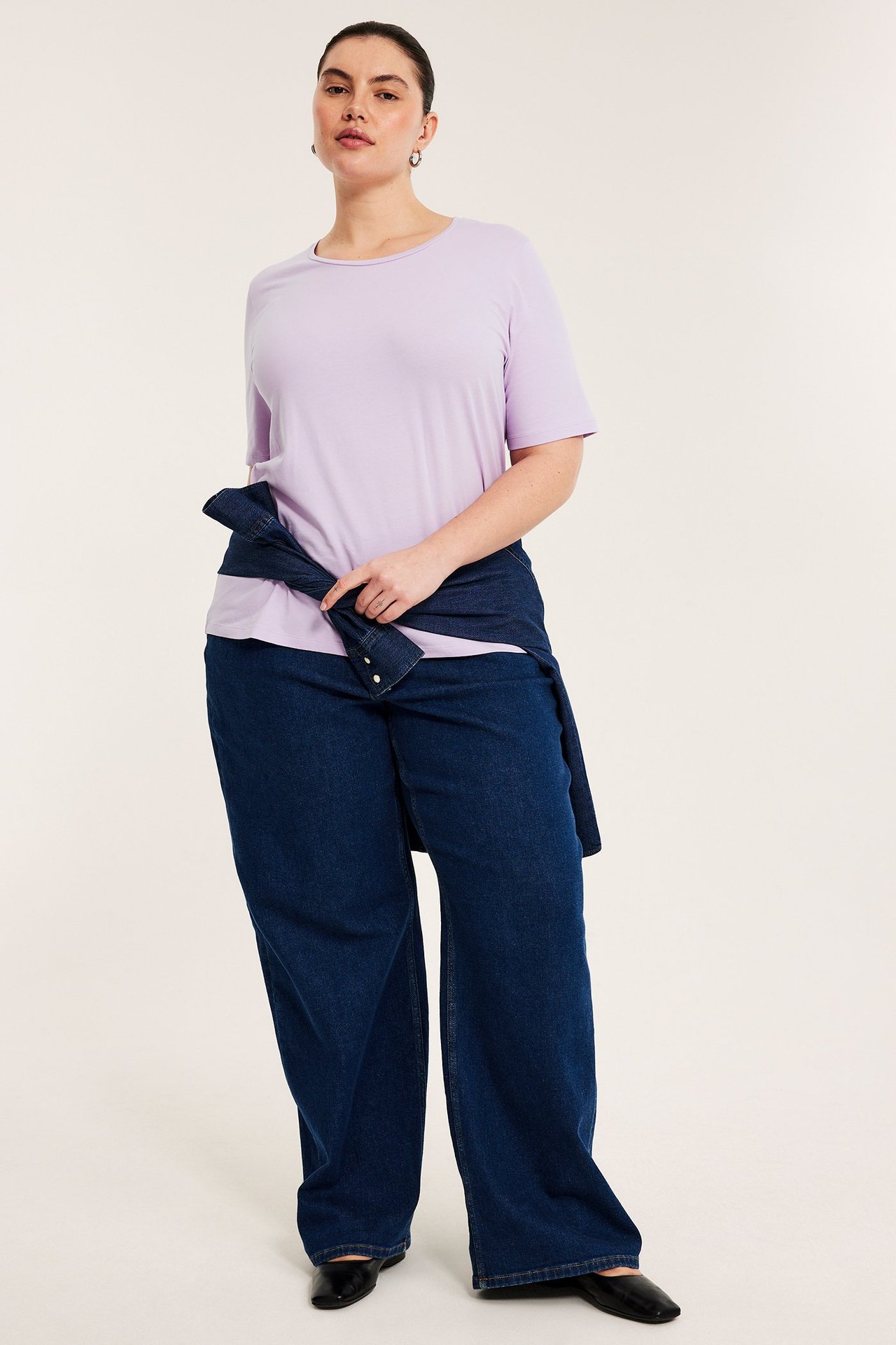 Widok z przodu: Kobieta w jasnofioletowym T-shircie, niebieskich szerokich jeansach i dżinsowej koszuli zawiązanej w pasie.