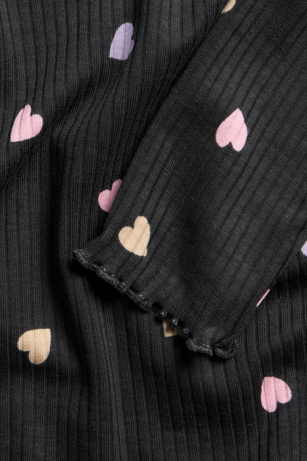 Nærbilde av svart ribbet langermet topp med rosa, beige og lys lilla hjertemønster og babylock-mansjetter.
