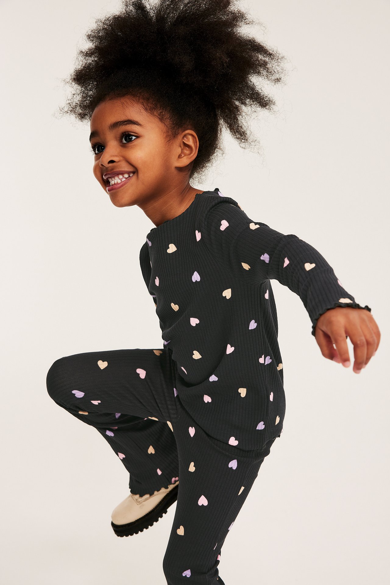 Forfra: Jente i svart ribbet langermet topp med fargerike hjertemønster og matchende utsvingte bukser.