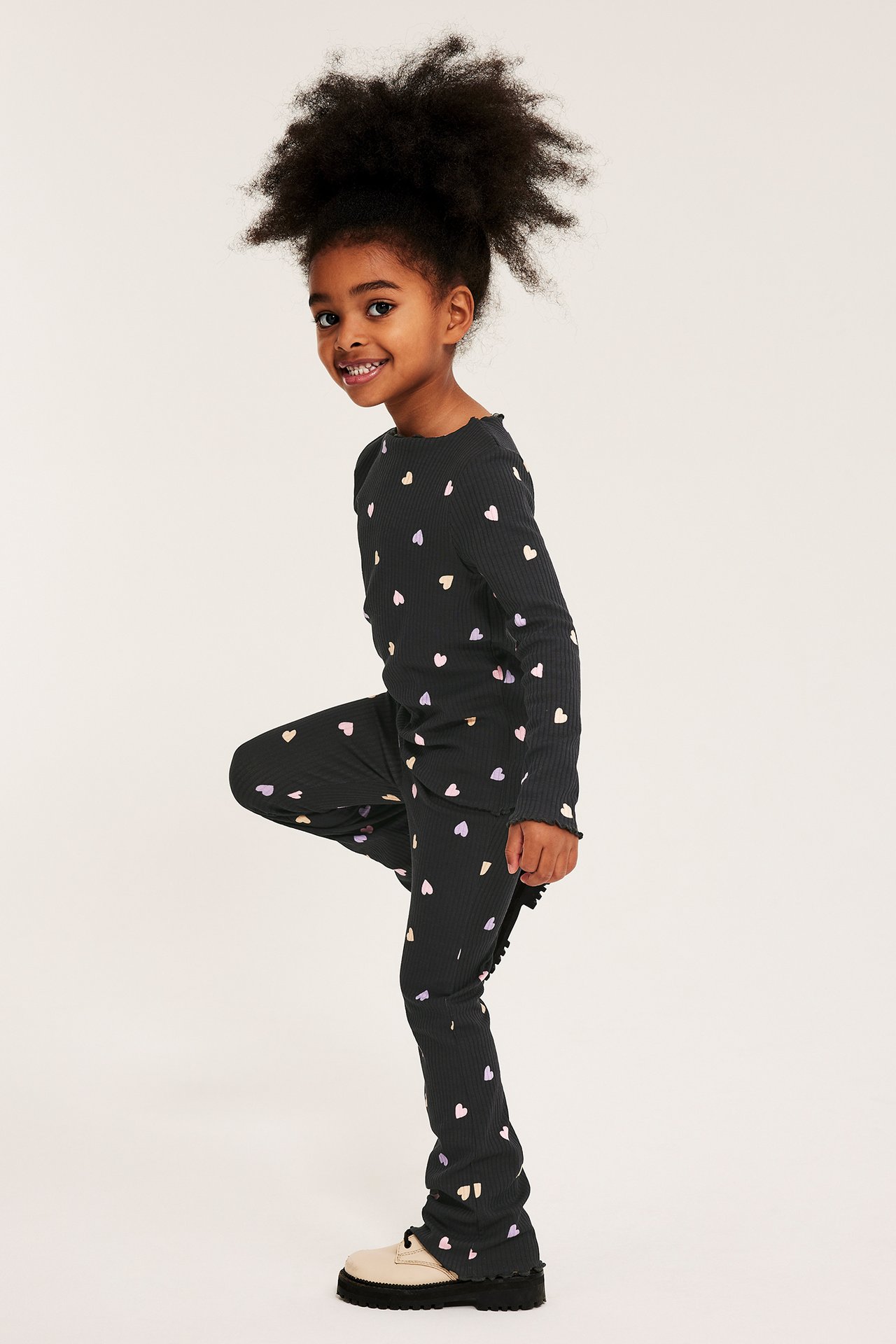 Sideutsikt av et barn i svarte ribbestrikkede flare leggings med hjertemønster, matchende topp og kremfargede sko.