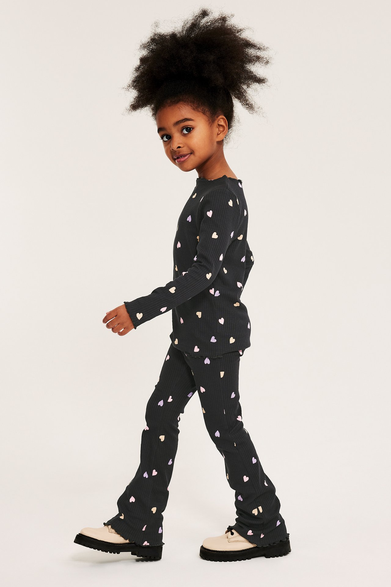 Sidevisning: Barn i svarte ribbestrikkede flare leggings og matchende topp med fargerike hjerte-mønster.