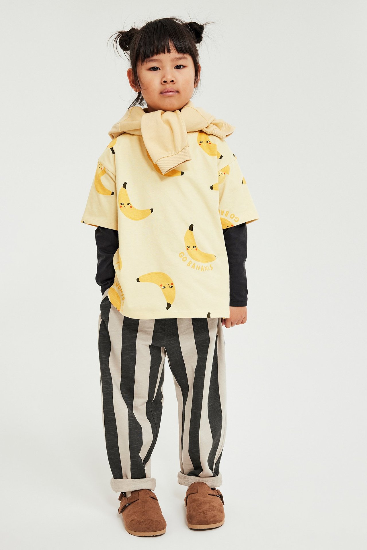 Framifrån: Barn i beige banantryckt t-shirt, svart långärmad tröja, beige hoodie, randiga byxor, bruna skor.