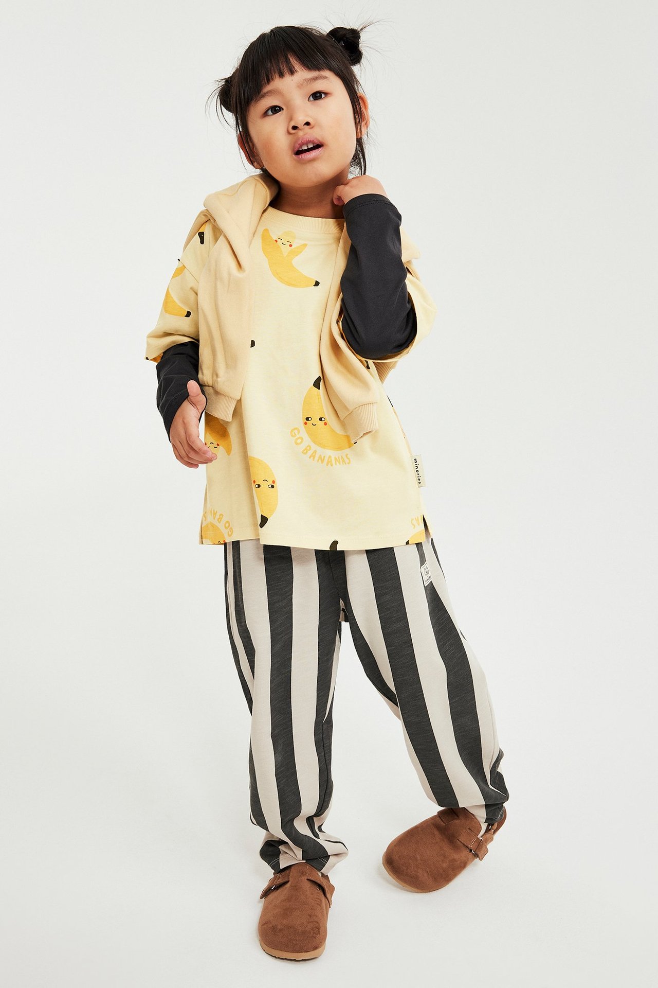 Framifrån: barn i beige bananmönstrad t-shirt över svart långärmad, randiga byxor och bruna tofflor.