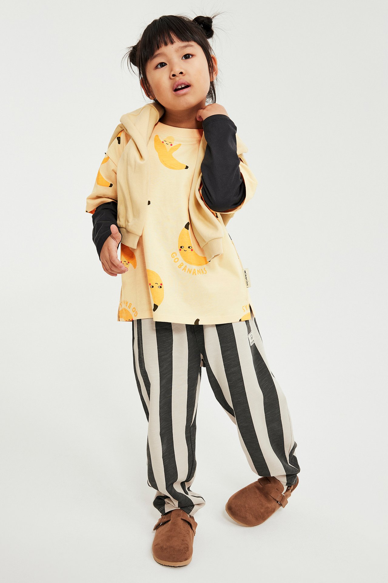 Widok z przodu dziecka w beżowym T-shircie w banany, czarnym longsleevie, pasiastych spodniach i brązowych chodakach.