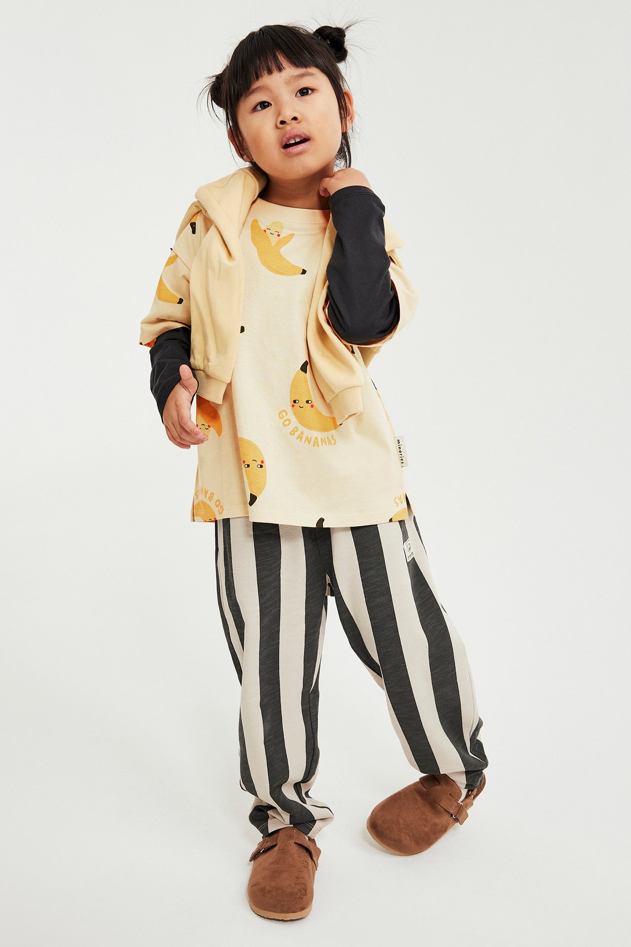 Lapsi yllään beige banaanipaita, musta pitkähihainen, beige huppari, raidalliset housut ja ruskeat puukengät.