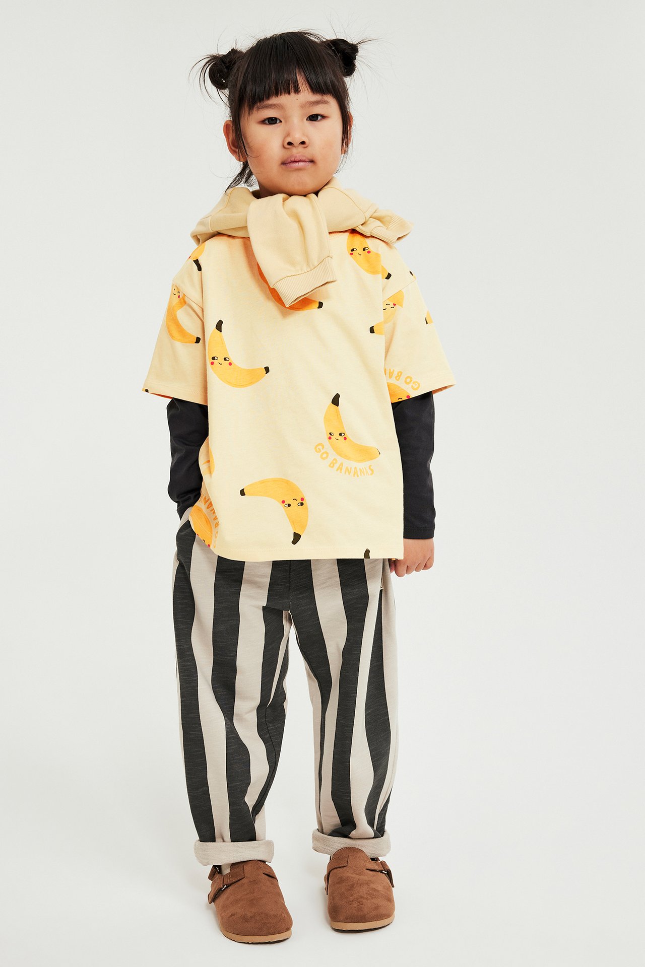 Barn bär beige t-shirt med banantryck, matchande huvtröja, svart långärmad, randiga byxor, bruna träskor.
