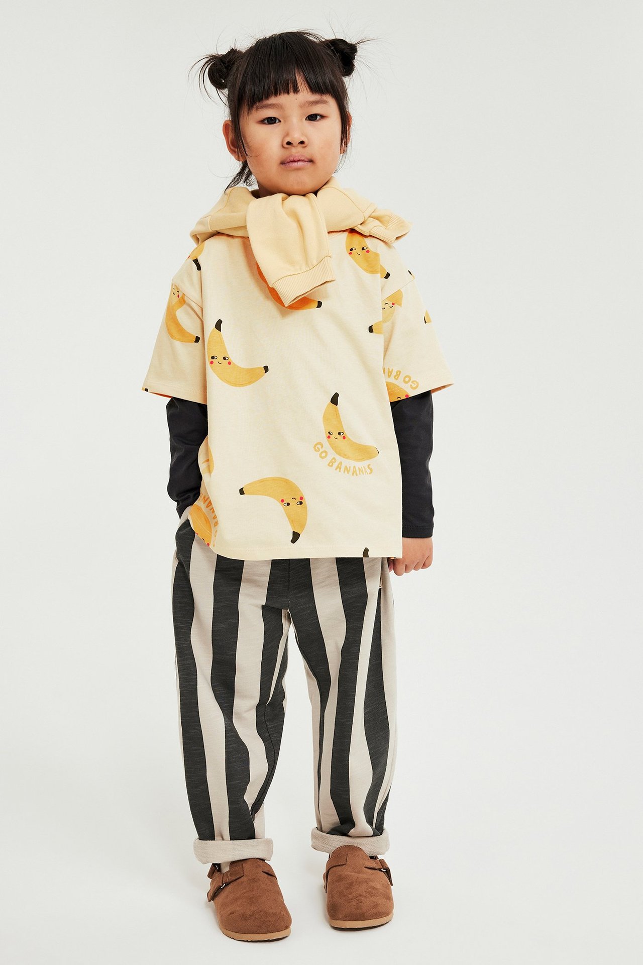 Lapsi beigessä banaanikuvioidussa t-paidassa, musta pitkähihainen, raidalliset housut, ruskeat kengät, keltainen huppari.