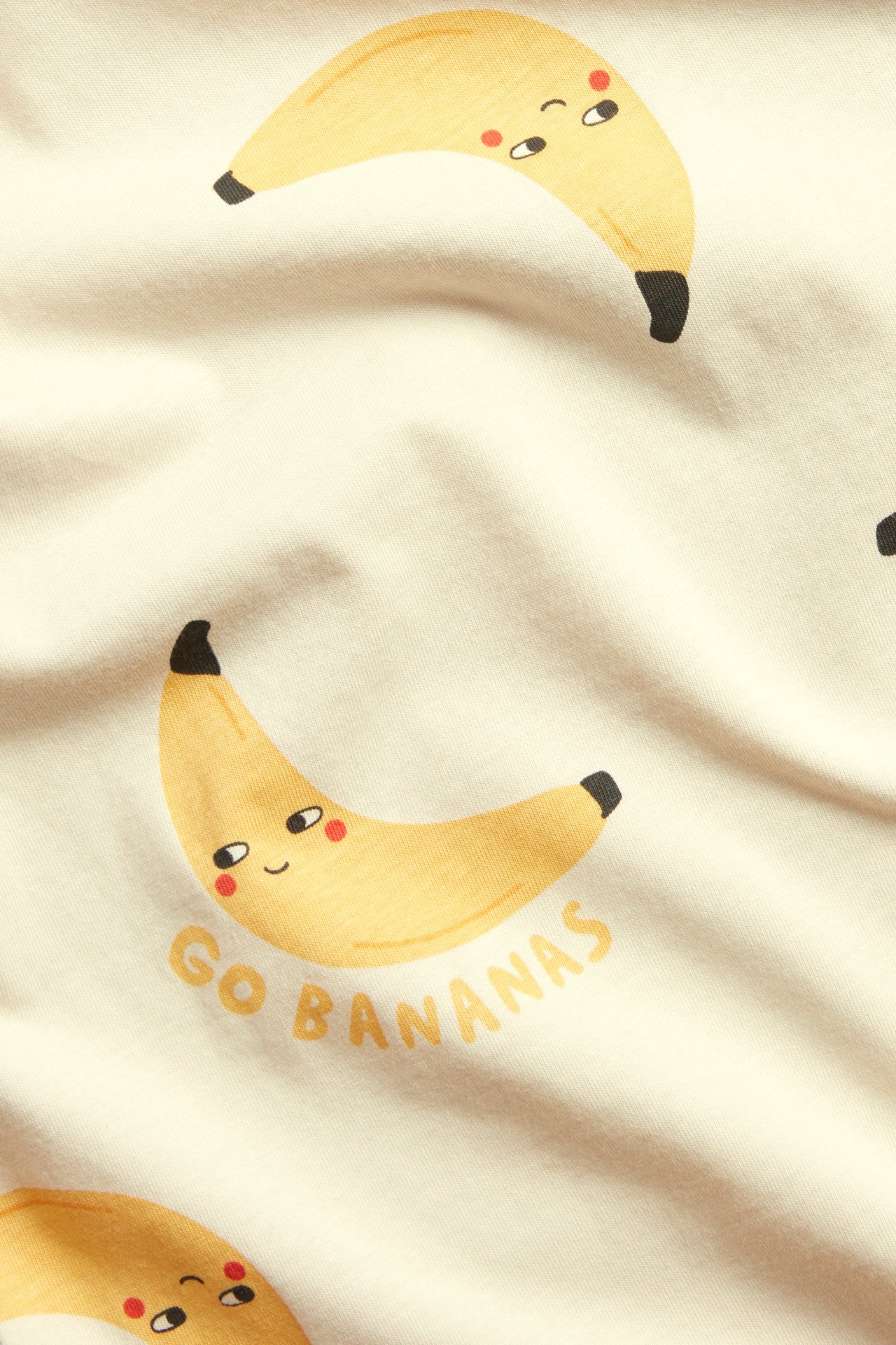 Närbild av en beige bomulls t-shirt med gula bananer med ansikten och texten 'Go Bananas'.