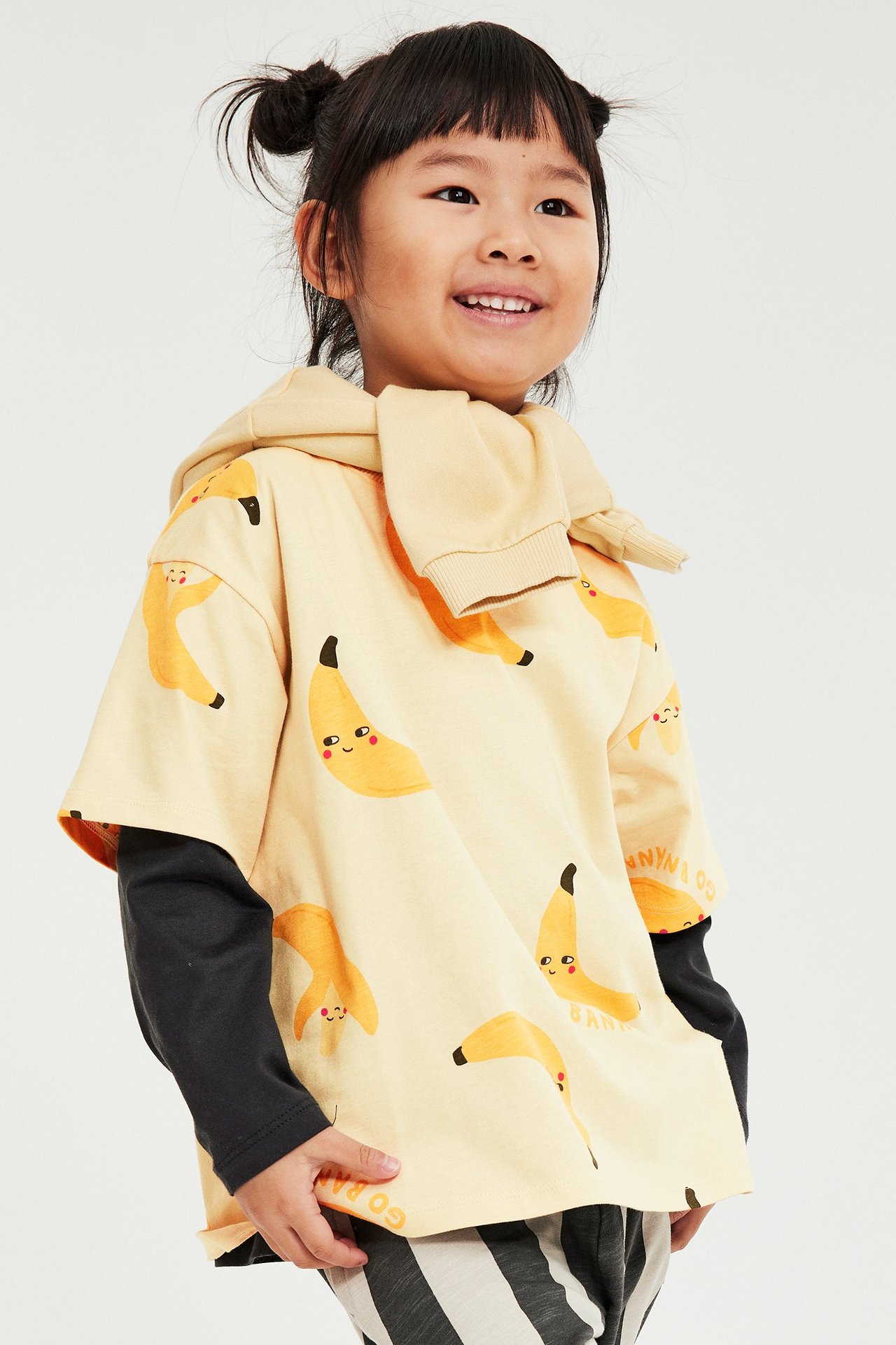 Framifrån: Barn i beige t-shirt med banantryck, svart långärmad topp, randiga byxor, beige huvtröja.