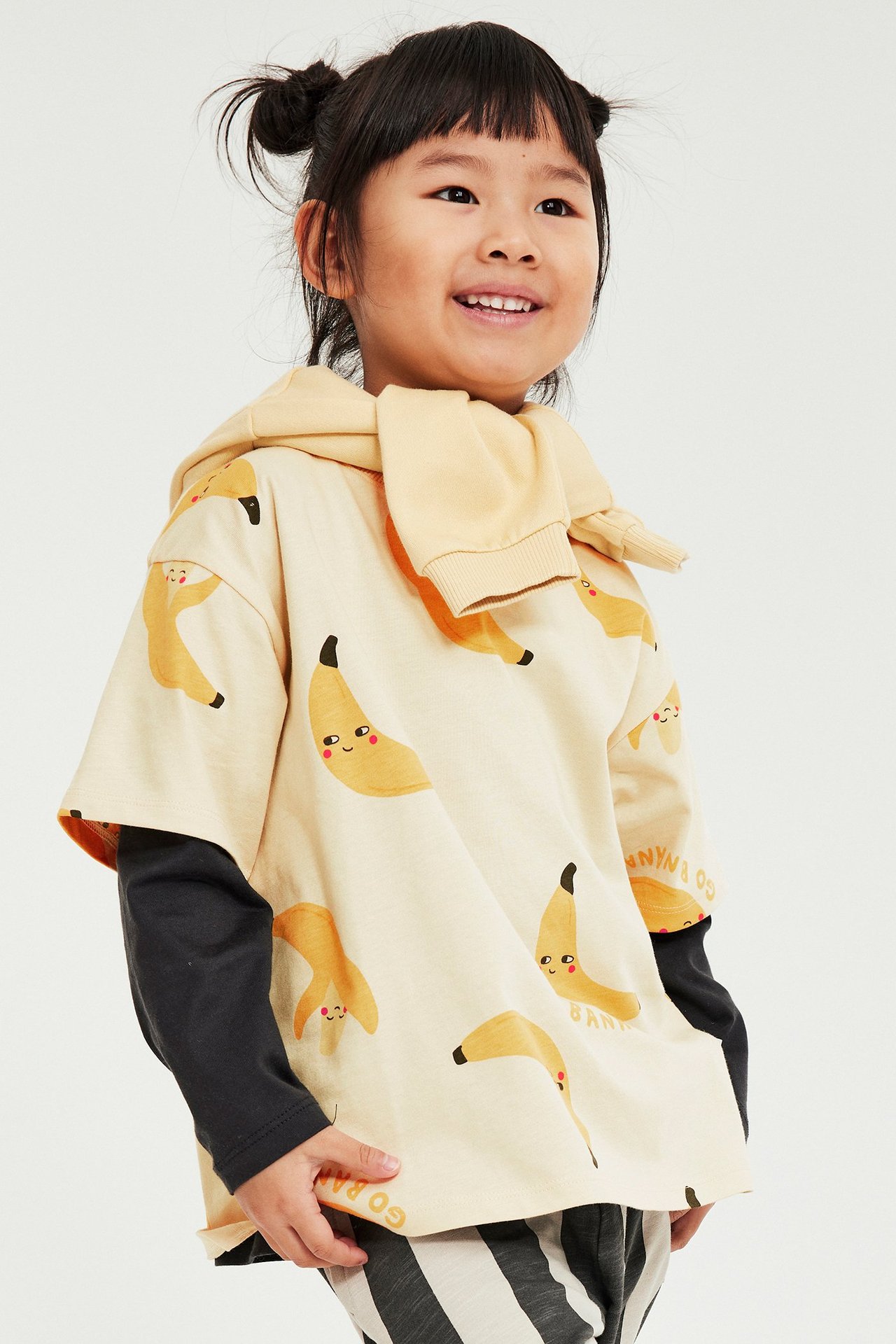 Framifrån: Ett barn i beige bomulls-t-shirt med banantryck över svart långärmad tröja och randiga byxor.