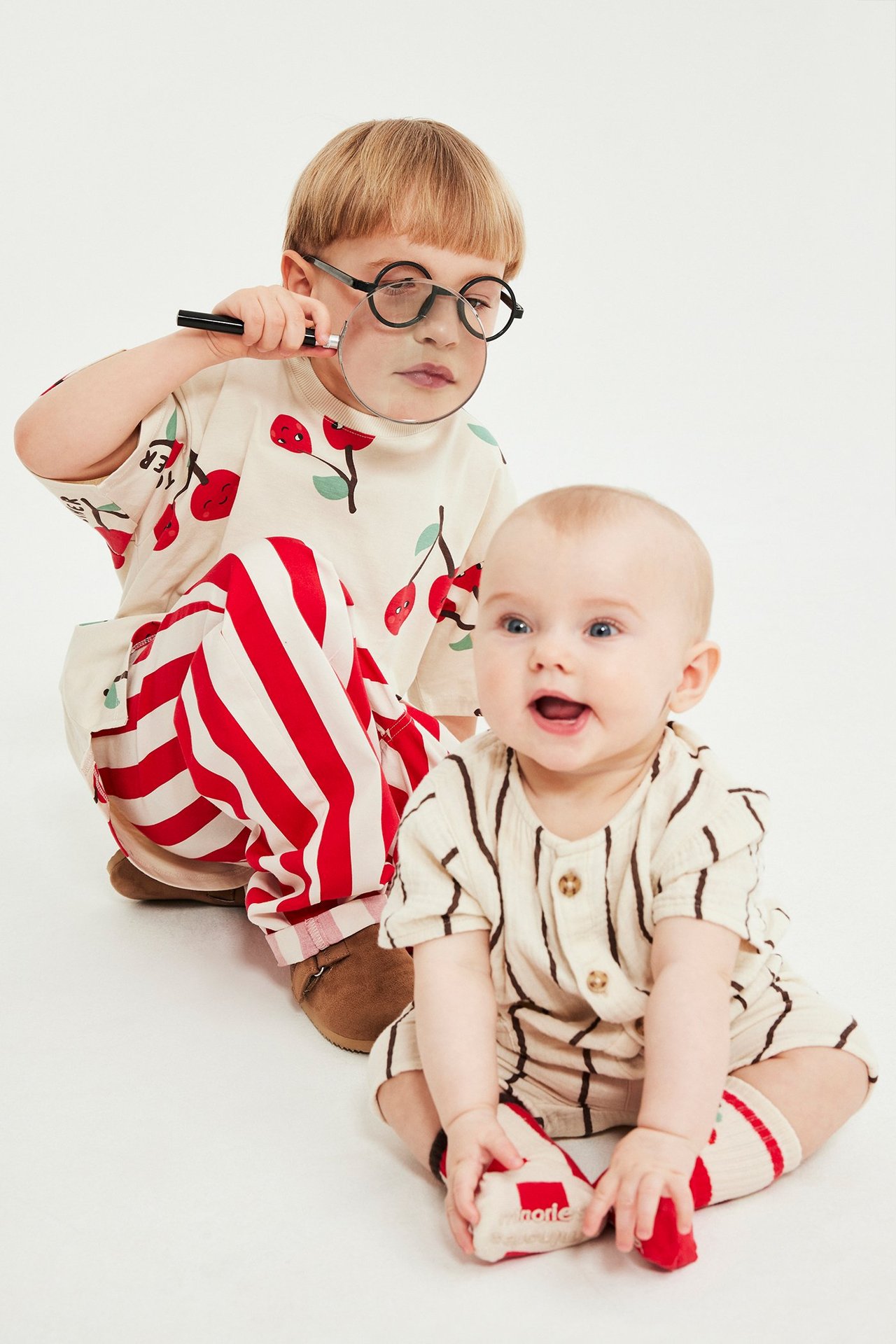 Przód: Dziecko w beżowej koszulce z czerwonym nadrukiem owoców, w pasiaste spodnie, obok niemowlę w pasiastej piżamce.