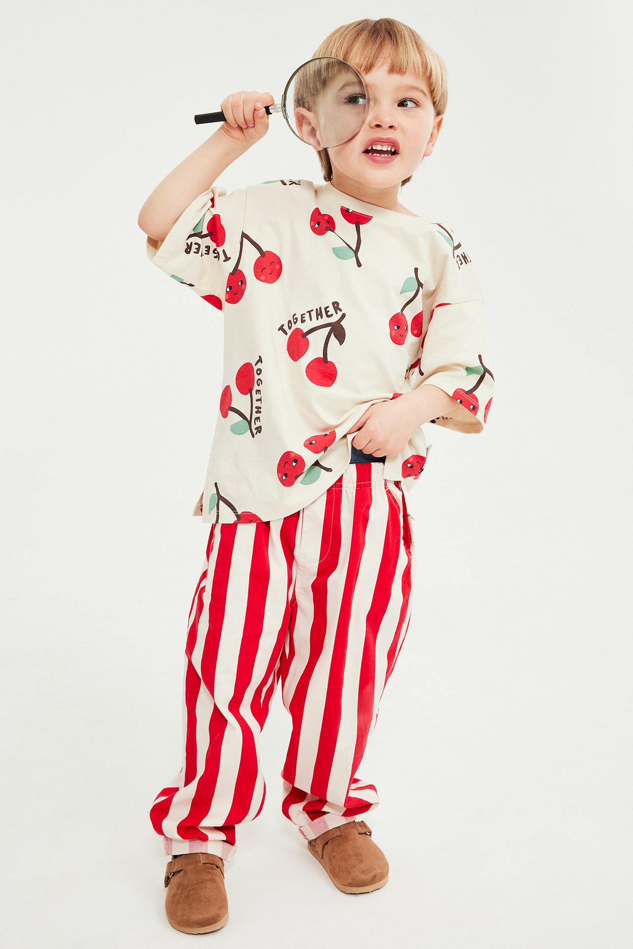 Barn i benvit körsbärsmönstrad T-shirt, randiga byxor, bruna skor, håller förstoringsglas. Framifrån.