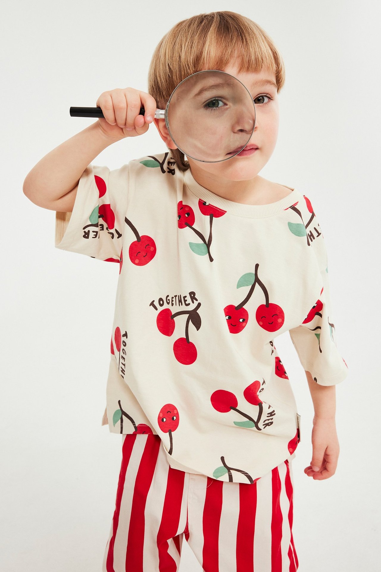 Barn bär offwhite t-shirt med rött körsbärstryck, röd-vitrandiga byxor, håller förstoringsglas.