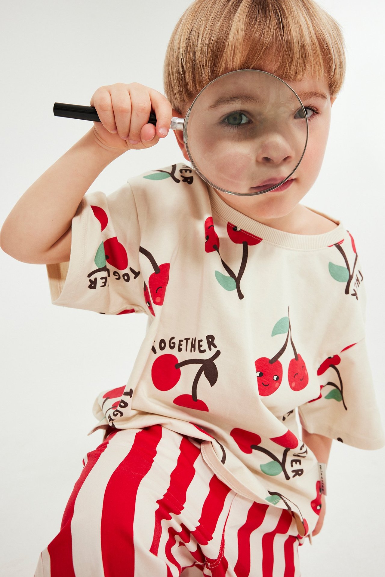 Widok z przodu: dziecko w kremowym t-shircie w wiśnie i czerwonych pasiastych szortach, trzymające lupę.
