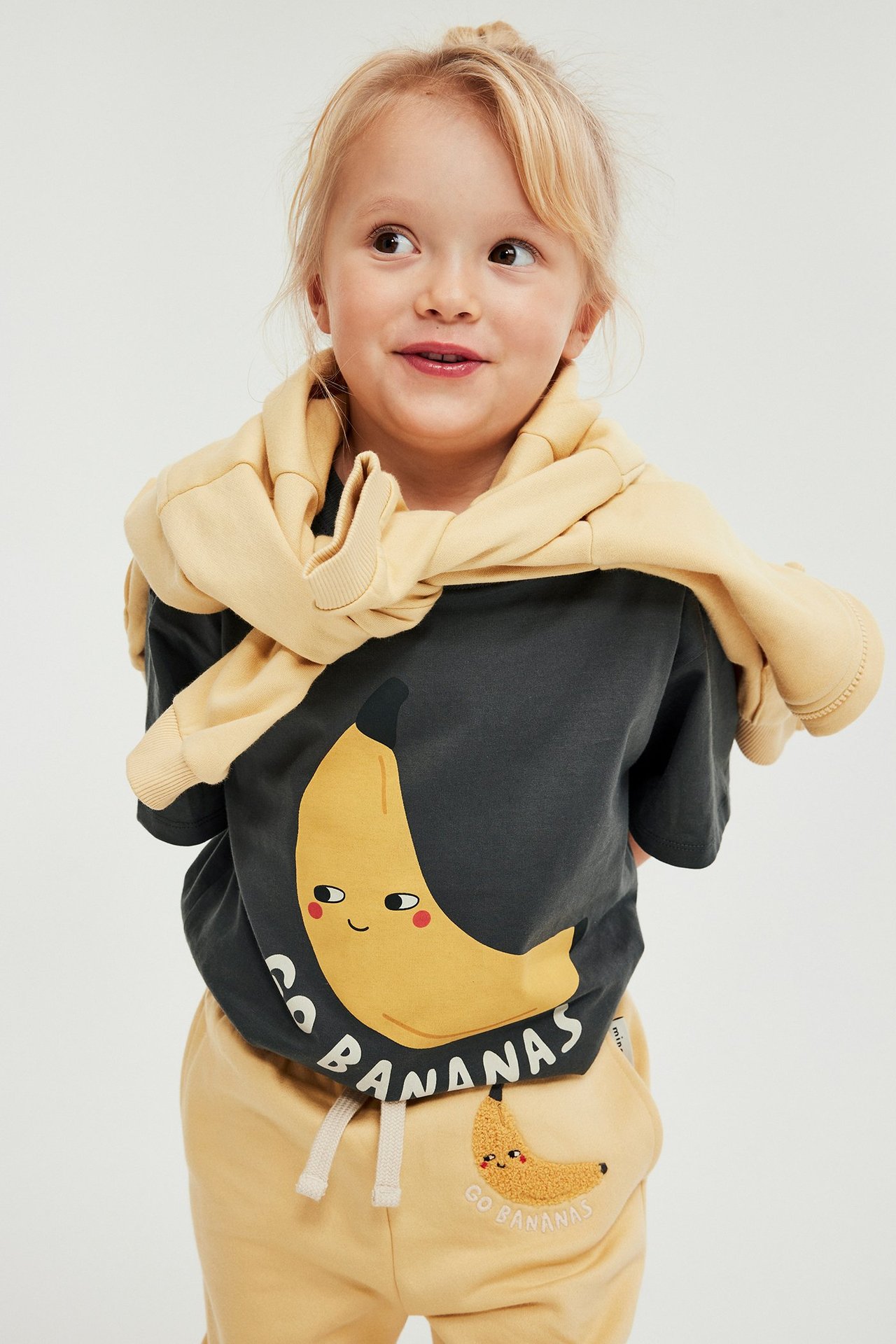 Lapsi edestä päin yllään musta t-paita banaanipainatuksella, keltaiset collegehousut ja keltainen collegepaita olkapäillä.