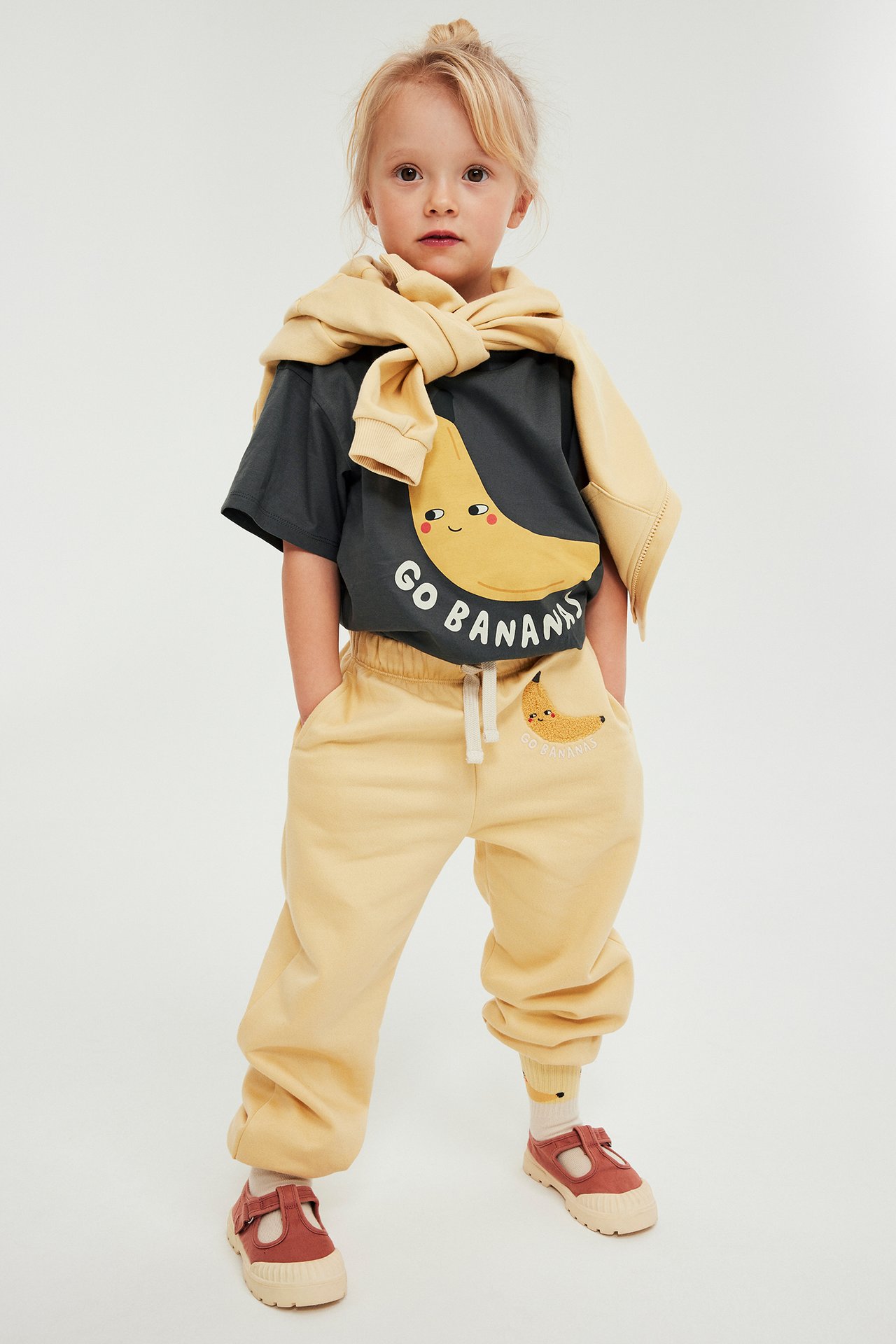 Frontvy: Barn bär mörkgrå t-shirt med gult banantryck, ljusgula sweatpants, matchande sweatshirt över axlarna.