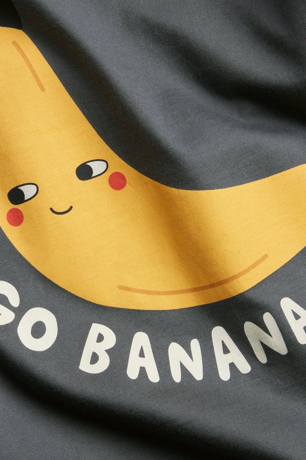 Zbliżenie czarnego bawełnianego T-shirtu z uśmiechniętym żółtym bananem i napisem 'GO BANANA'.