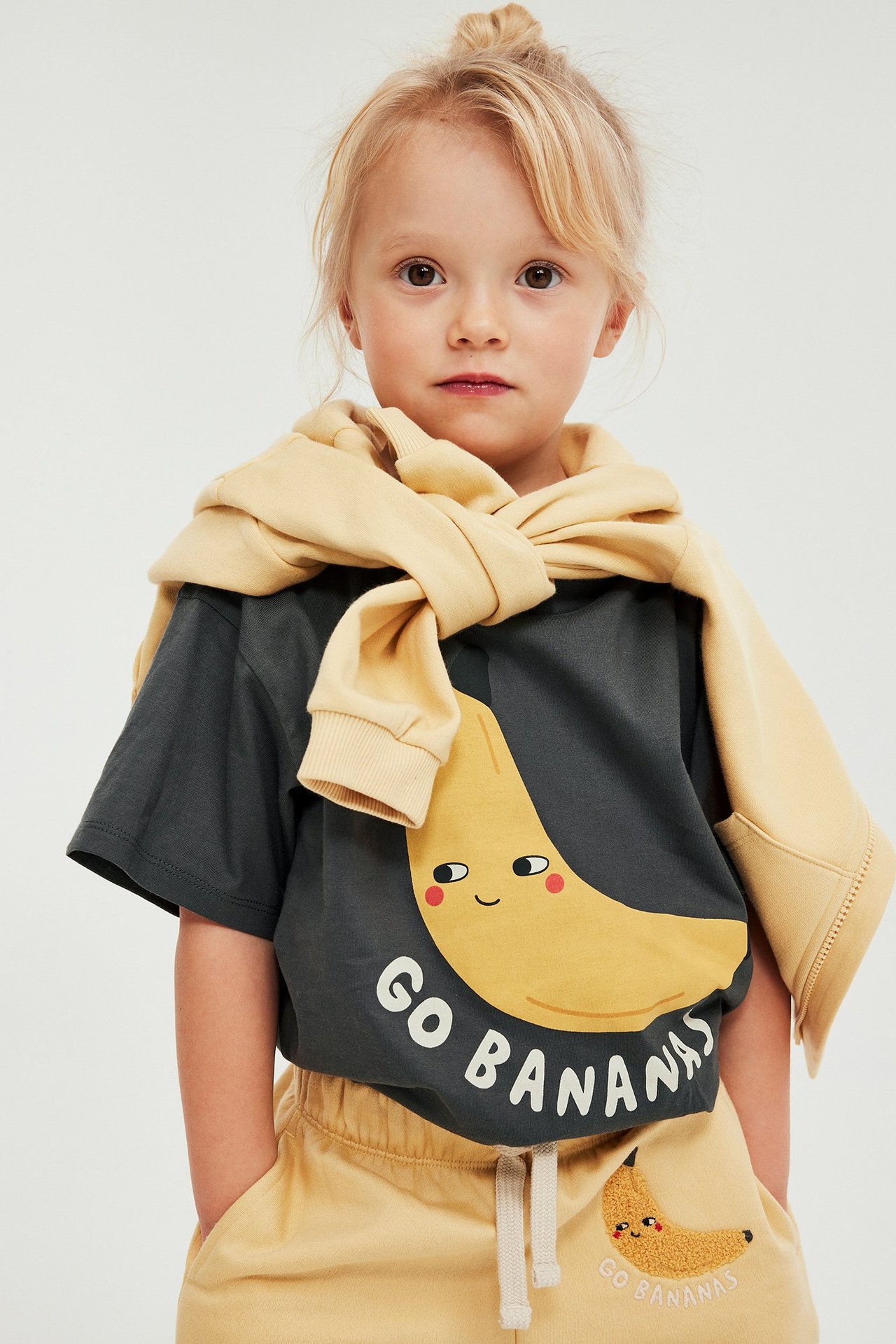 Edestä: lapsi tummanharmaassa banaaniprintti-T-paidassa, keltainen collegepaita ja keltaiset shortsit.