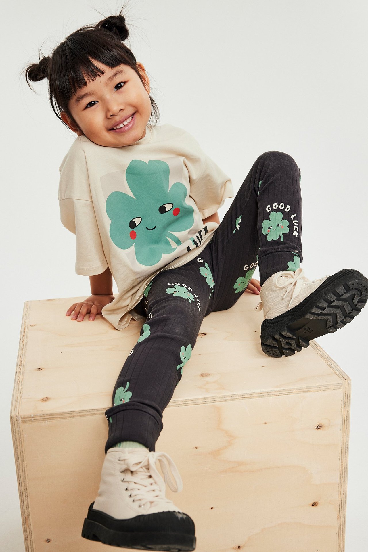 Widok z przodu dziecka w beżowym t-shircie z zielonym nadrukiem, ciemnoszarych legginsach i beżowych butach.