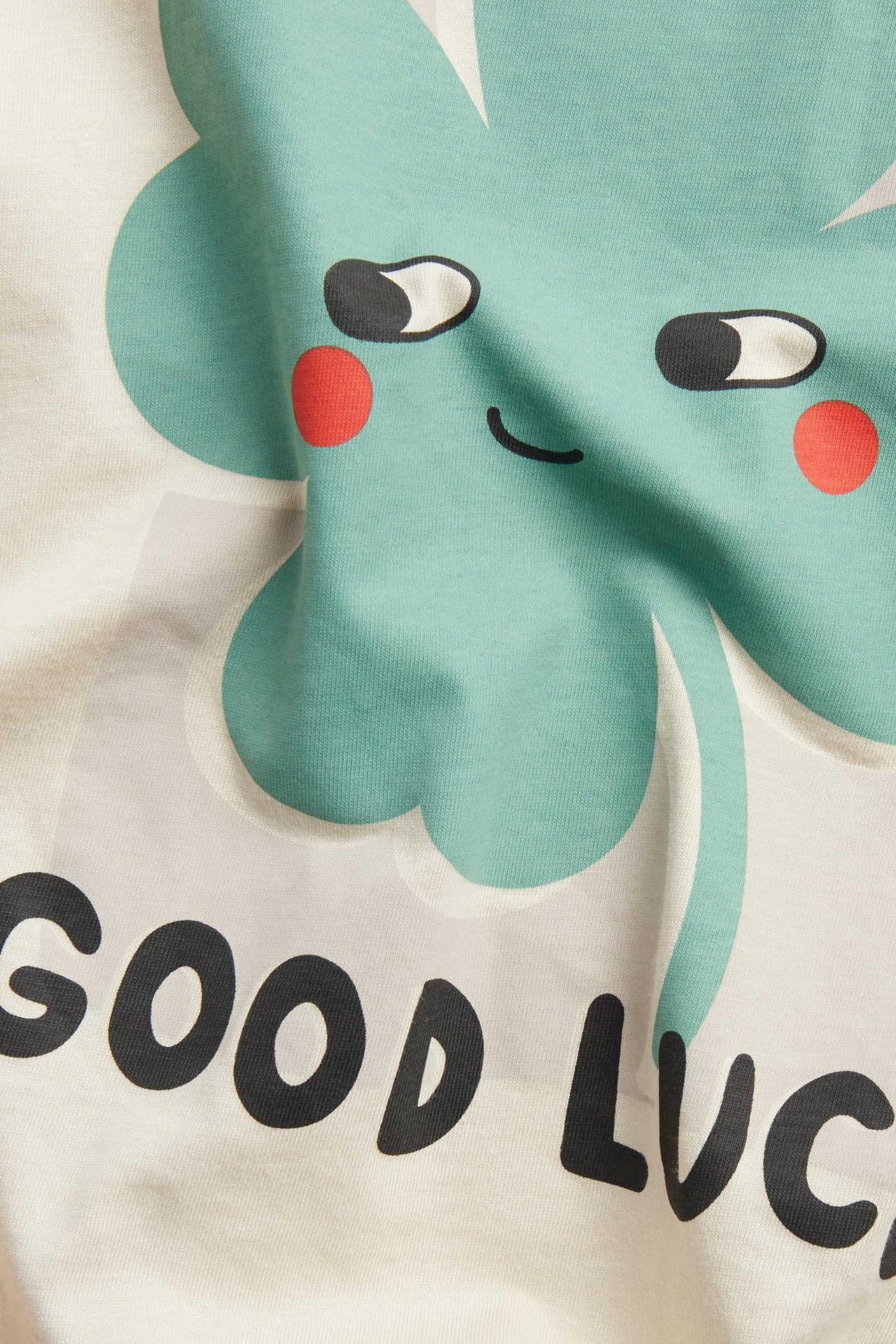 Zbliżenie beżowego bawełnianego T-shirtu z dużym jasnoturkusowym nadrukiem koniczyny i napisem „GOOD LUC”.