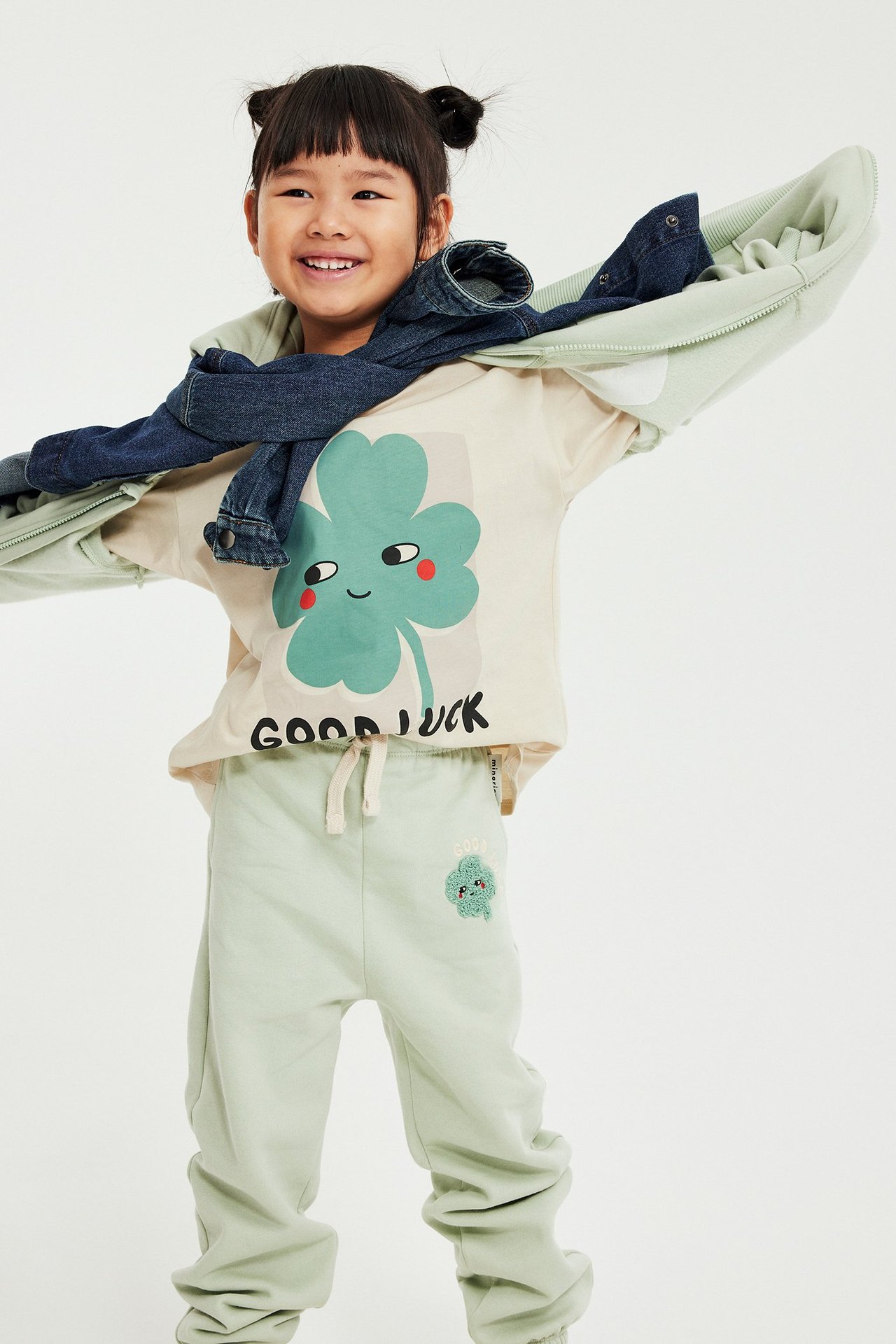 Widok z przodu: Dziecko w beżowej koszulce z zieloną koniczyną, jasnozielonych dresach i kurtce dżinsowej na ramionach.