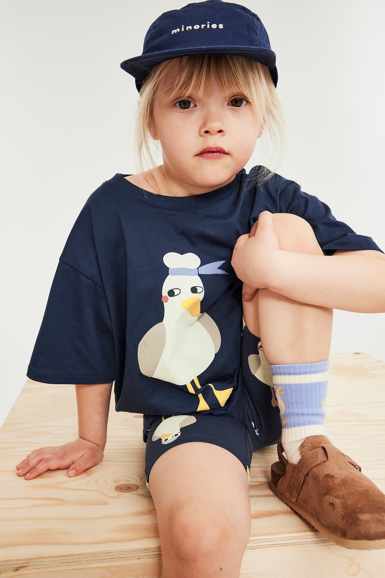Barn i blå t-shirt med måstryck, matchande shorts, blå keps, randiga strumpor och bruna tofflor.