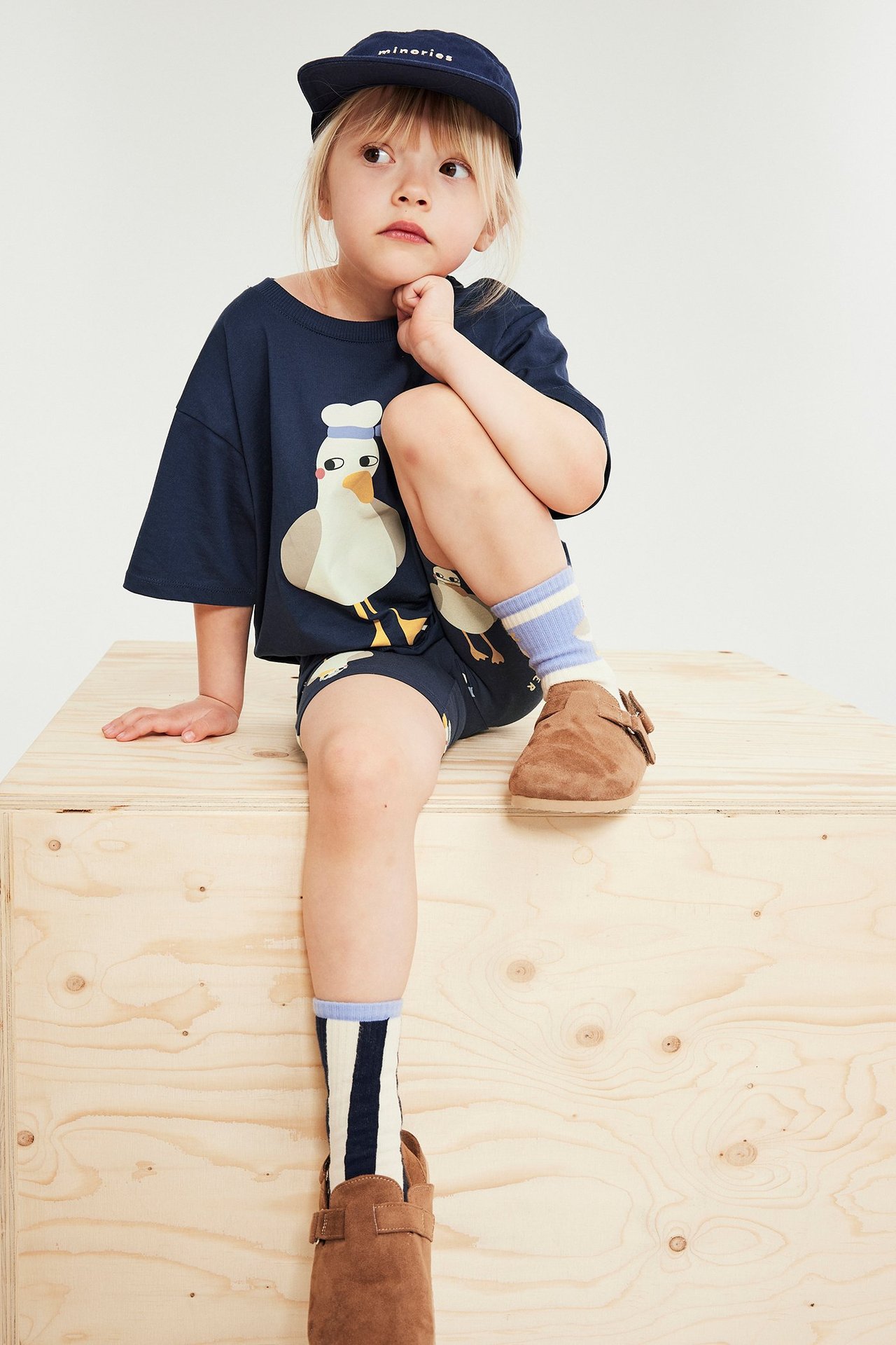 Barn bär mörkblå t-shirt med måstryck, mörka shorts, marinblå keps, randiga strumpor, bruna skor.