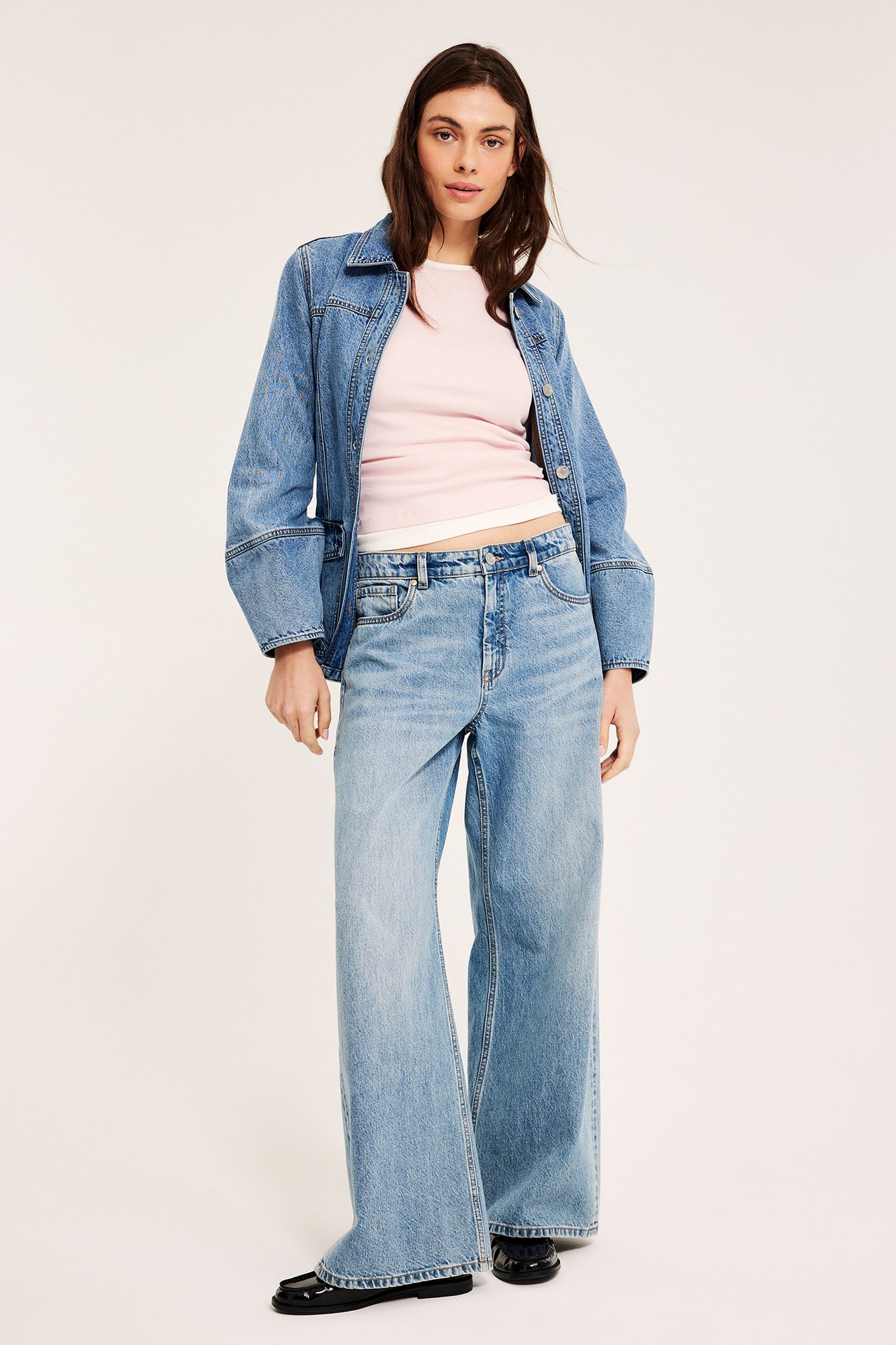 Forfra: kvinne i lyserosa ribbet langermet topp, blå dongerijakke og vide dongeribukser.