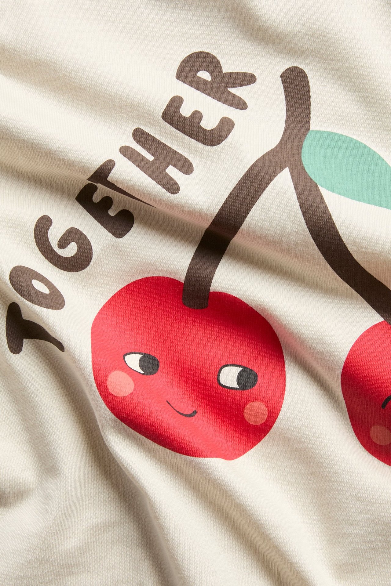 Närbild på ljusbeige t-shirt i bomull med tryck av leende röda körsbär och texten 'TOGETHER'.
