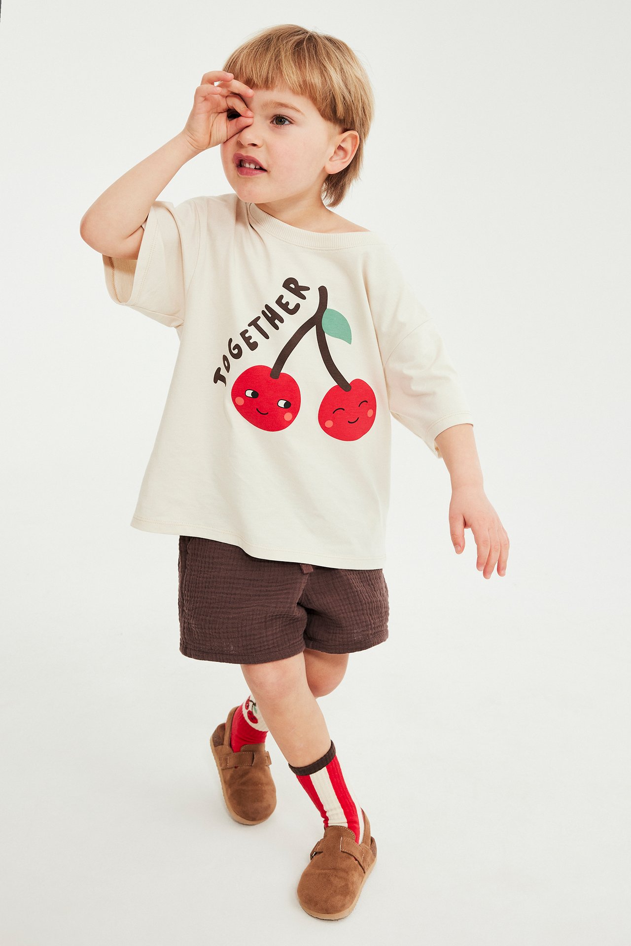 Lapsi edestäpäin yllään vaalea beige kirsikkaprintti-t-paita, ruskeat shortsit, raidalliset sukat, ruskeat kengät.