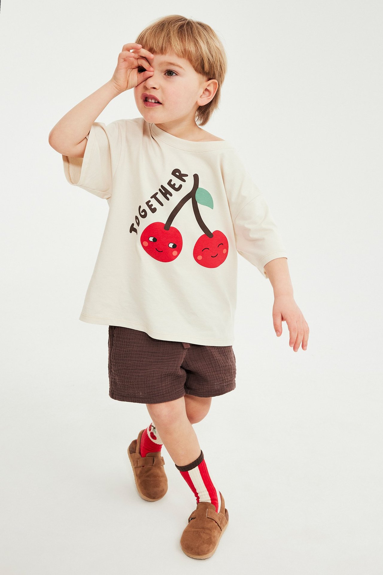 Lapsi edestäpäin yllään vaalea beige kirsikkaprintti-t-paita, ruskeat shortsit, raidalliset sukat, ruskeat kengät.