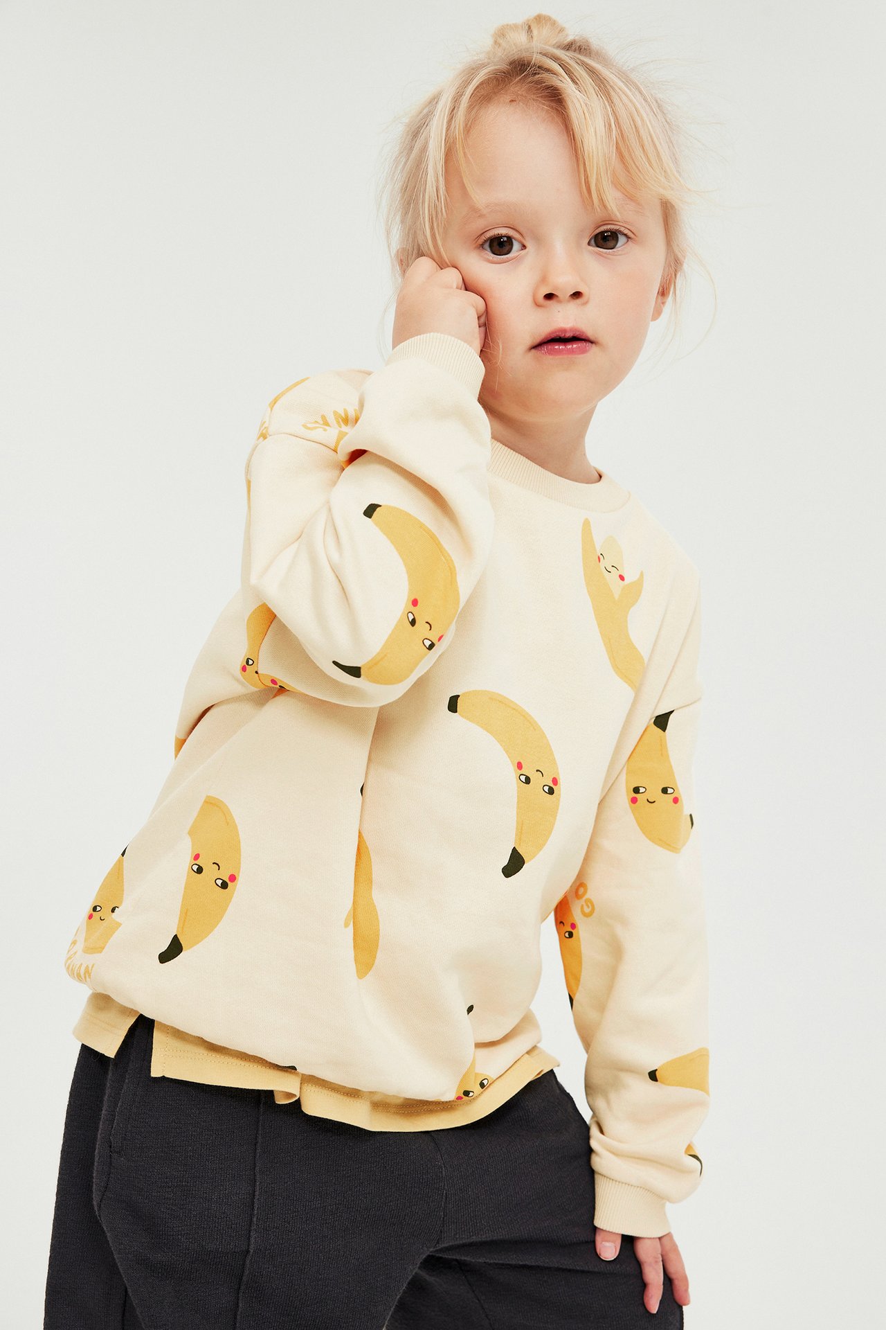 Edestäpäin: lapsi yllään beige pusero banaaniprintillä ja tummanharmaat collegehousut.