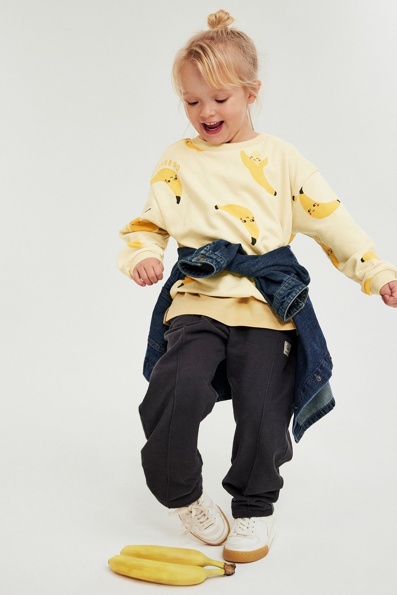 Barn i beige sweatshirt med bananmönster, mörkgrå mjukisbyxor, blå jeansjacka i midjan och vita sneakers.