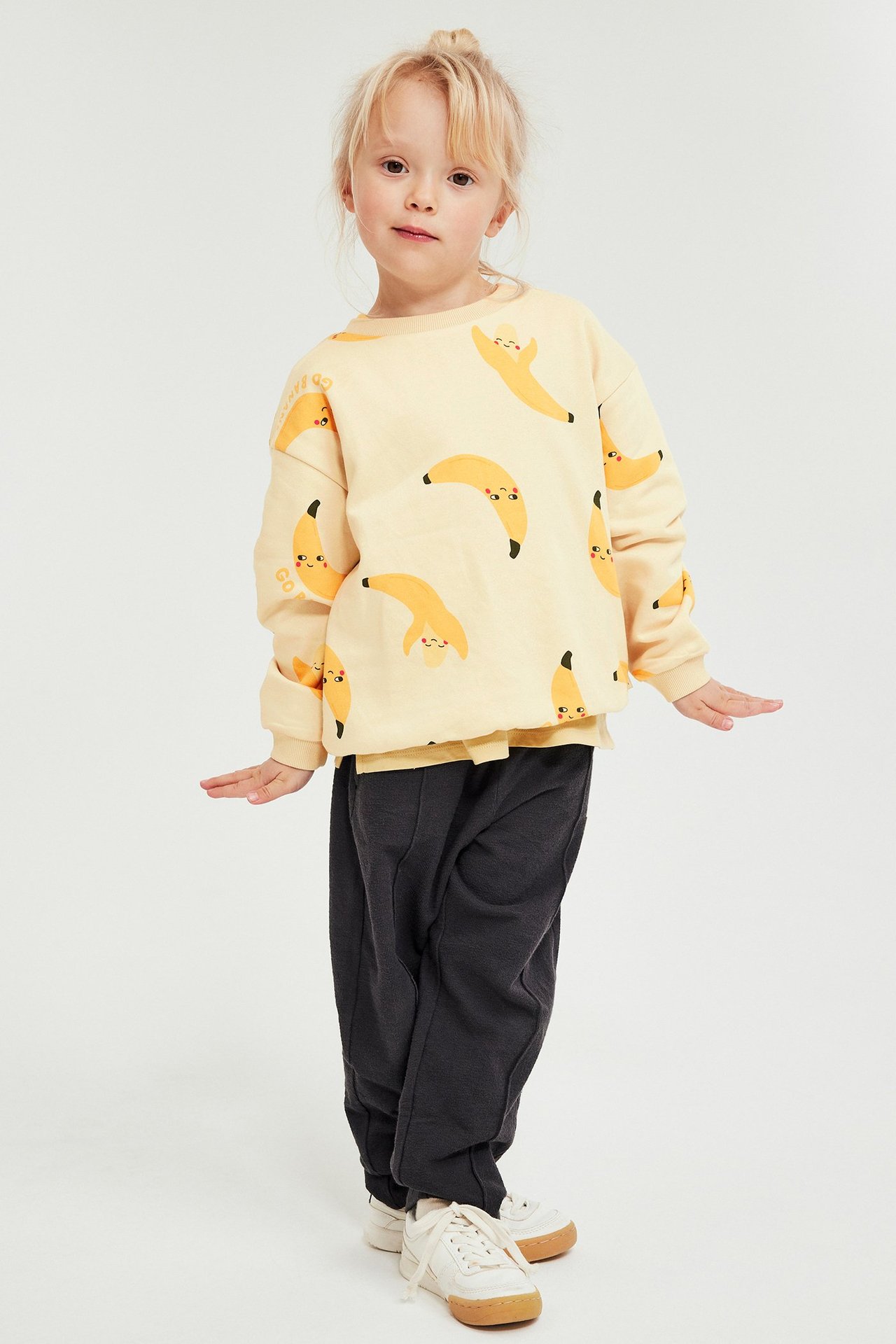 Forfra, et barn i en beige sweatshirt med bananprint, mørkegrå sweatpants og hvite joggesko.